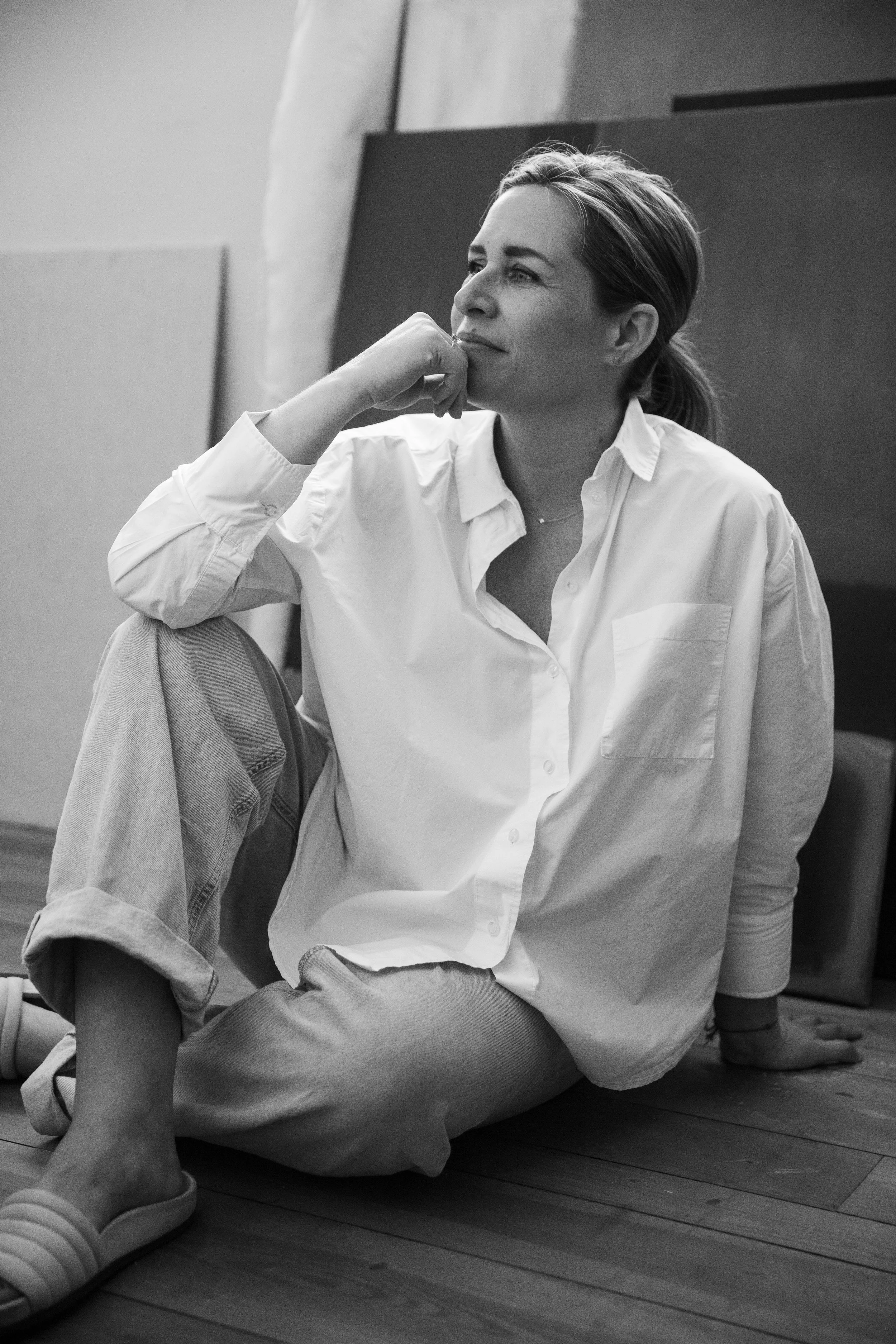 A woman sitting on the wooden floor with one knee up, wearing a white button-up shirt and jeans, looking thoughtfully to the side in a room with abstract paintings.