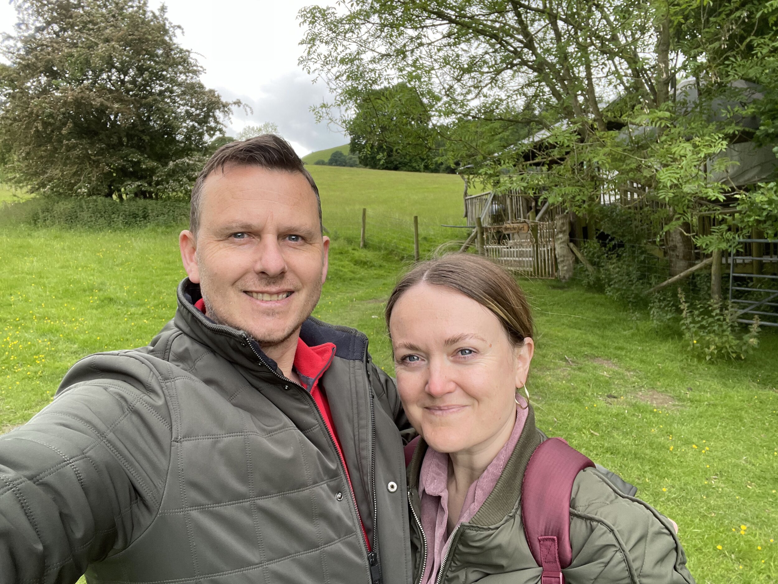 A man and a woman taking a selfie outdoors in a green, rural area with trees and a wooden fence in the background.