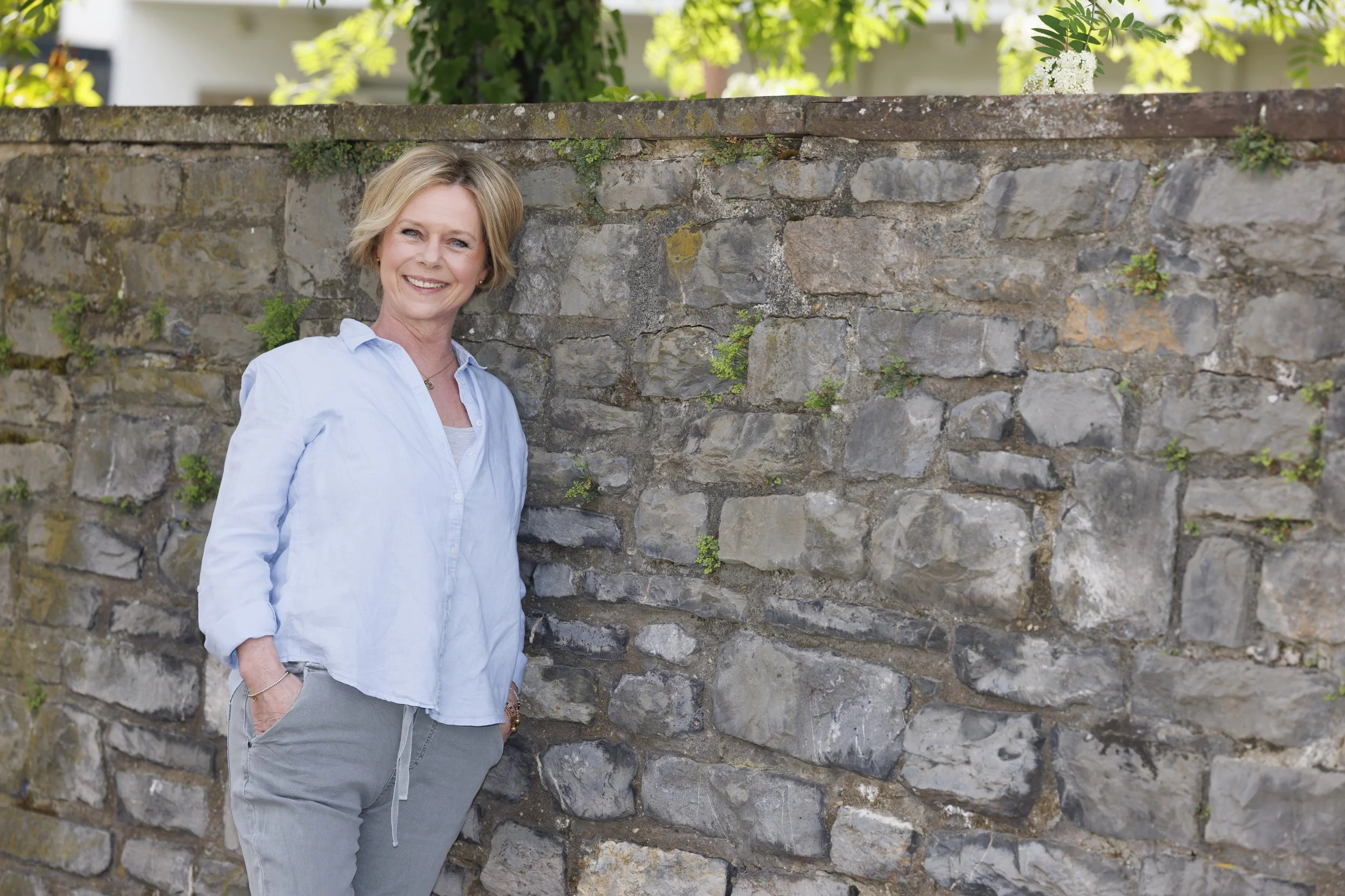 Eine lächelnde Frau steht an einer Steinmauer mit grünen Pflanzen und Blüten im Hintergrund.