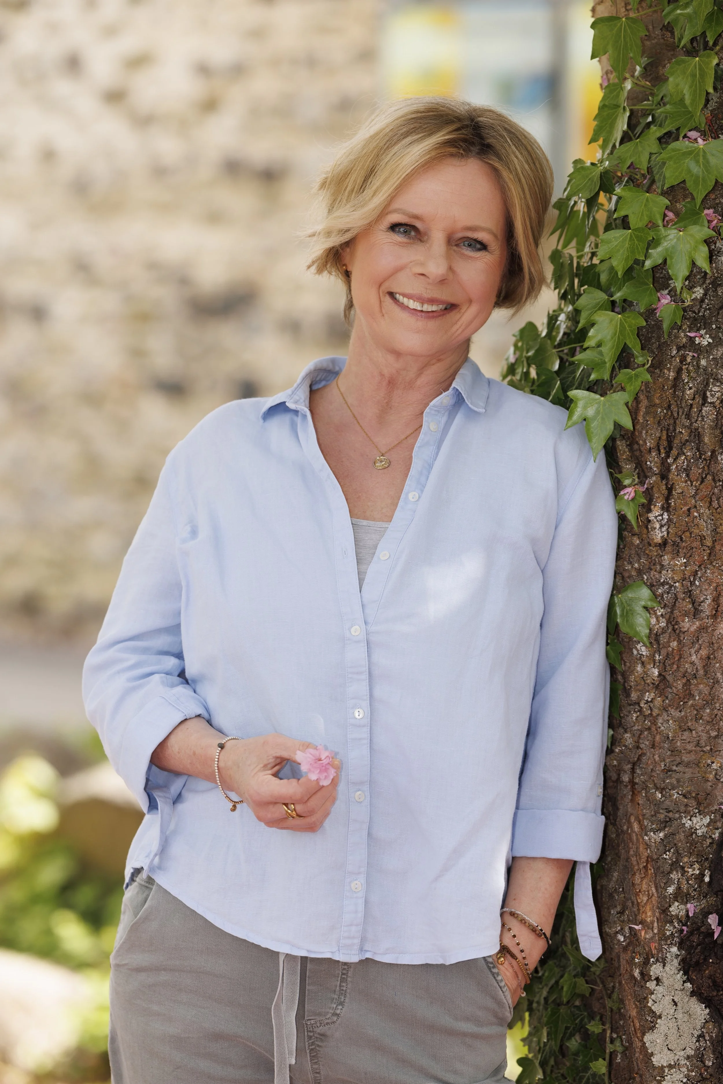 Eine lachende Frau mit kurzen blonden Haaren, die eine hellblaue Bluse trägt, steht neben einem Baum mit grünen Blättern und lila Blumen. Sie hält eine rosa Blume in der Hand und trägt Schmuck wie Halskette, Armbänder und Ringe. Der Hintergrund ist unscharf und zeigt ein helles, sonniges Außensetting.