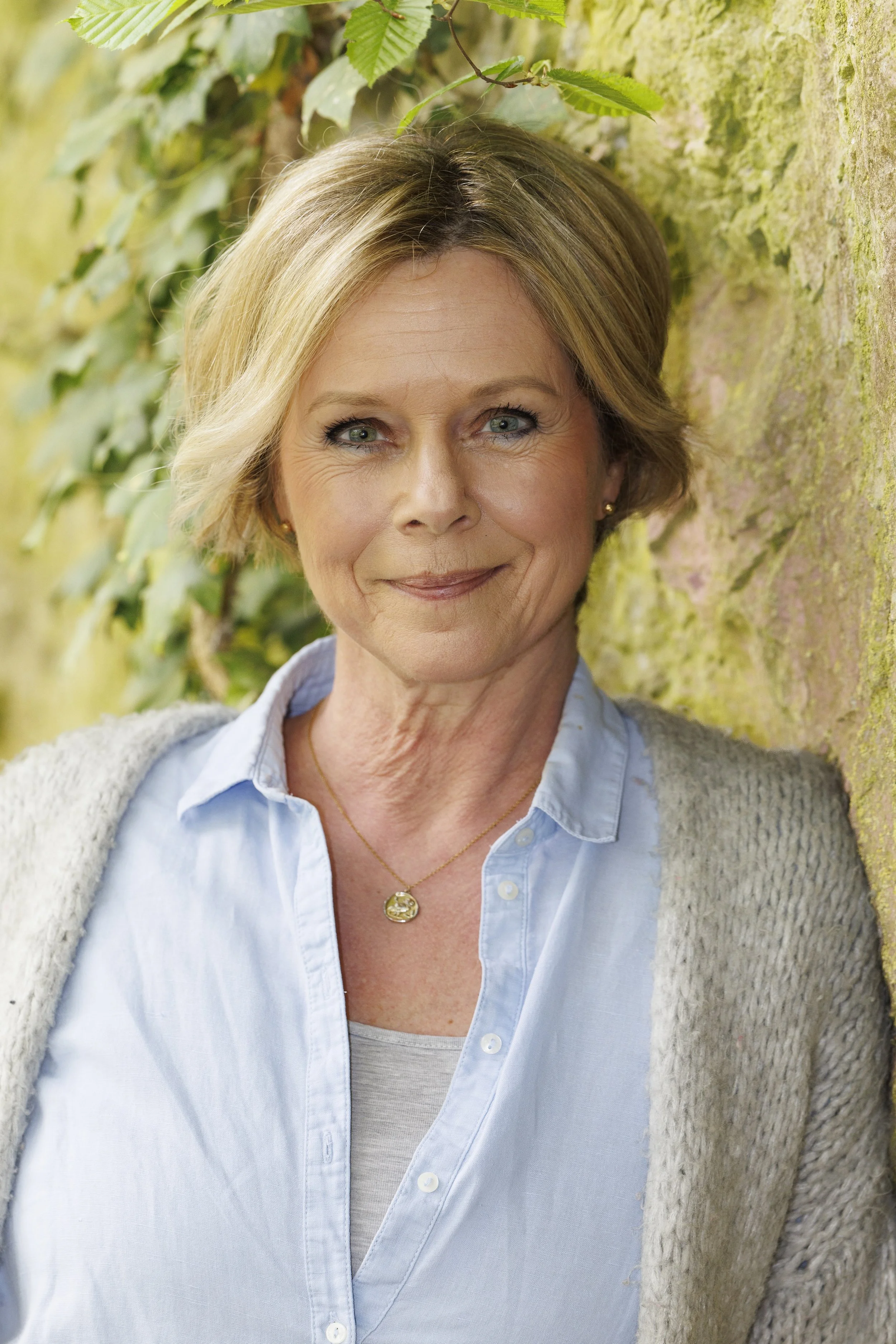 Porträt einer lächelnden, mittelalten Frau mit kurzen, blond gesträhnten Haaren, die an einer bewachsenen Steinmauer lehnt, trägt eine blaue Bluse, einen weißen cardigan und eine goldene Halskette, bei natürlichem Licht im Freien.