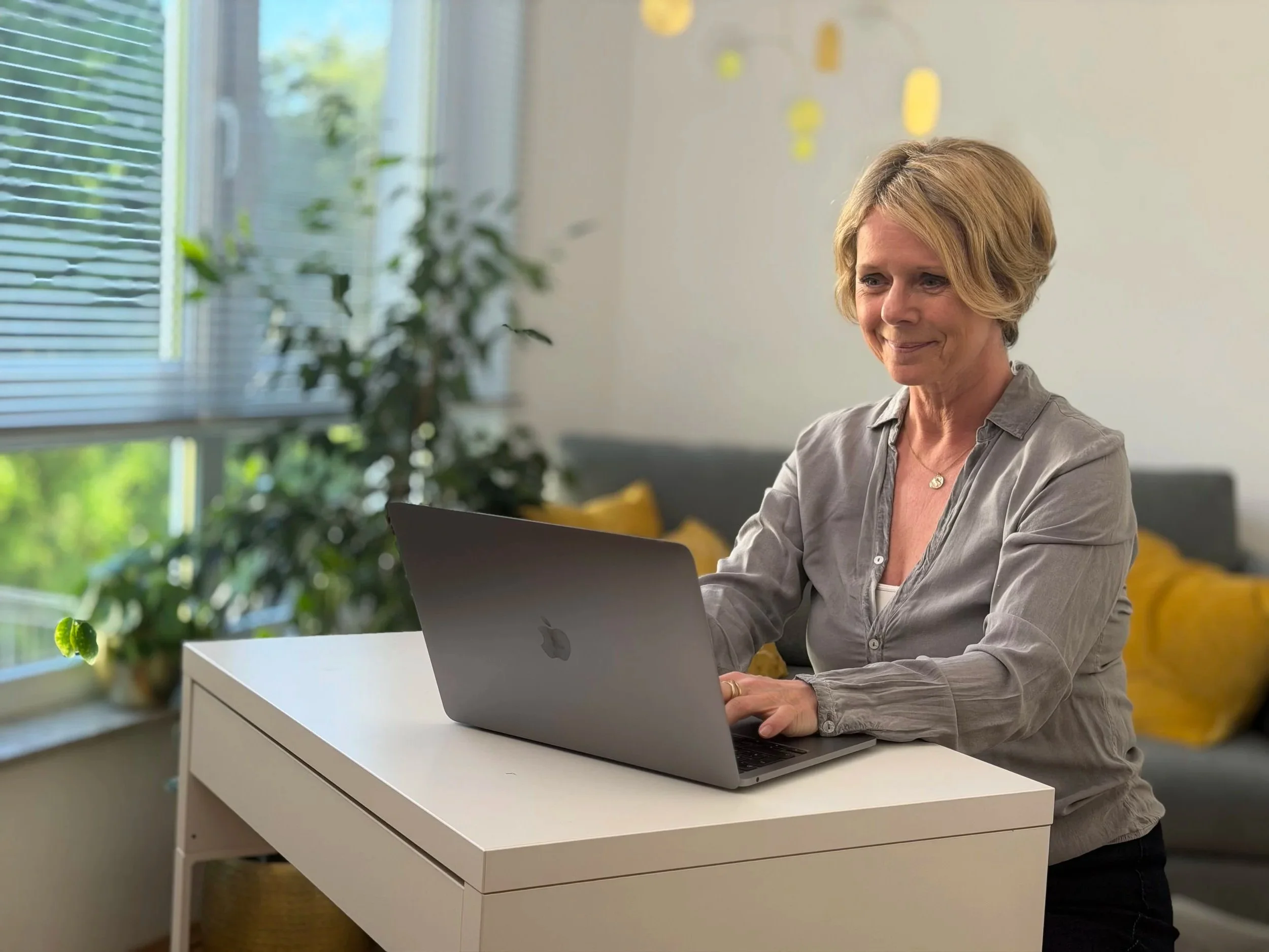 Eine Frau sitzt an einem weißen Tisch mit einem Laptop vor einem großen Fenster mit durchsichtigen Jalousien. Im Hintergrund sind grüne Pflanzen und gelbe Kissen auf einem Sofa zu sehen.