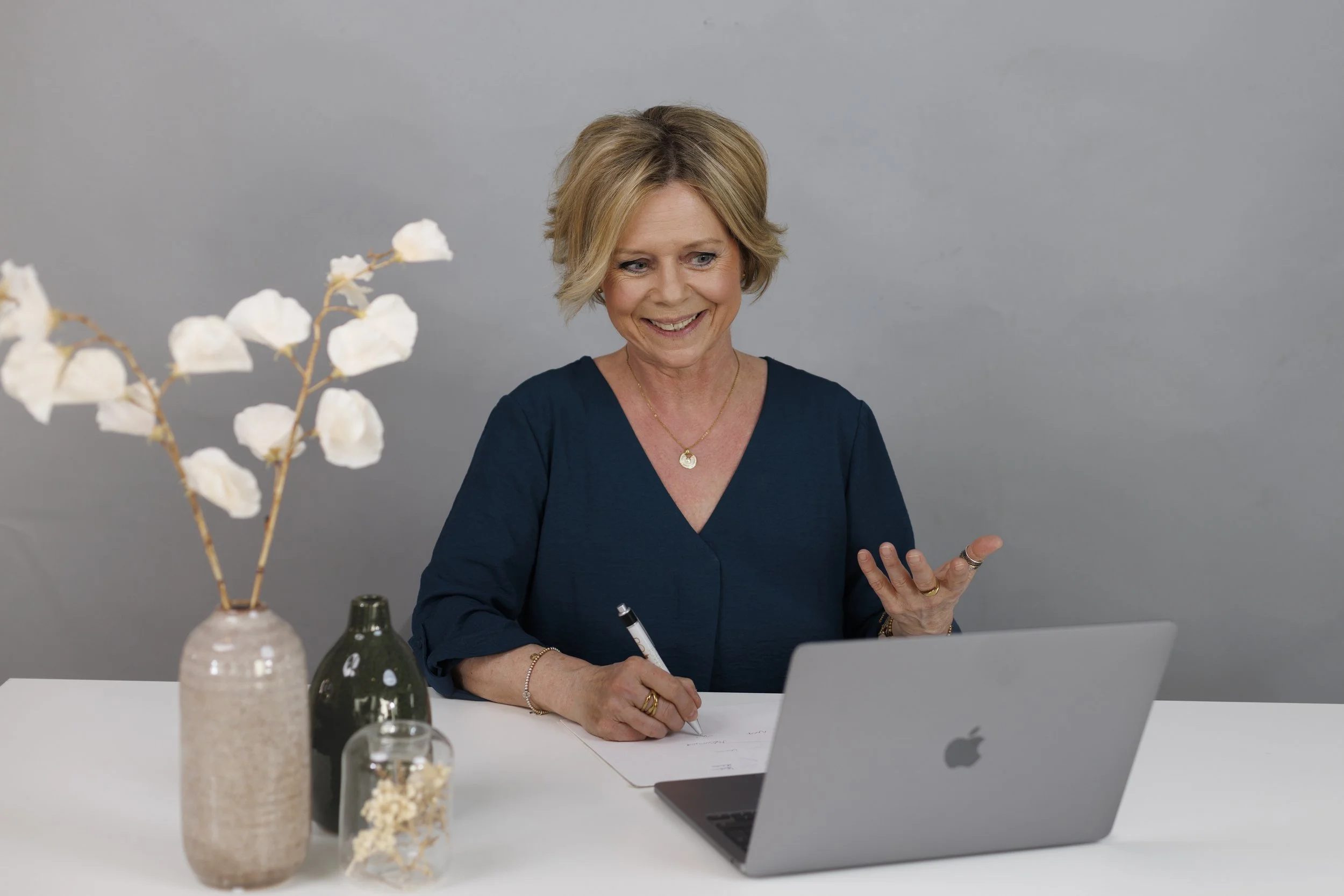 Eine lächelnde Frau mit kurzen blonden Haaren sitzt an einem weißen Tisch vor einem Laptop, während sie schreibt. Vor ihr stehen mehrere Vasen mit weißen und schwarzen Blumen. Sie trägt eine dunkle Bluse und eine goldene Halskette.