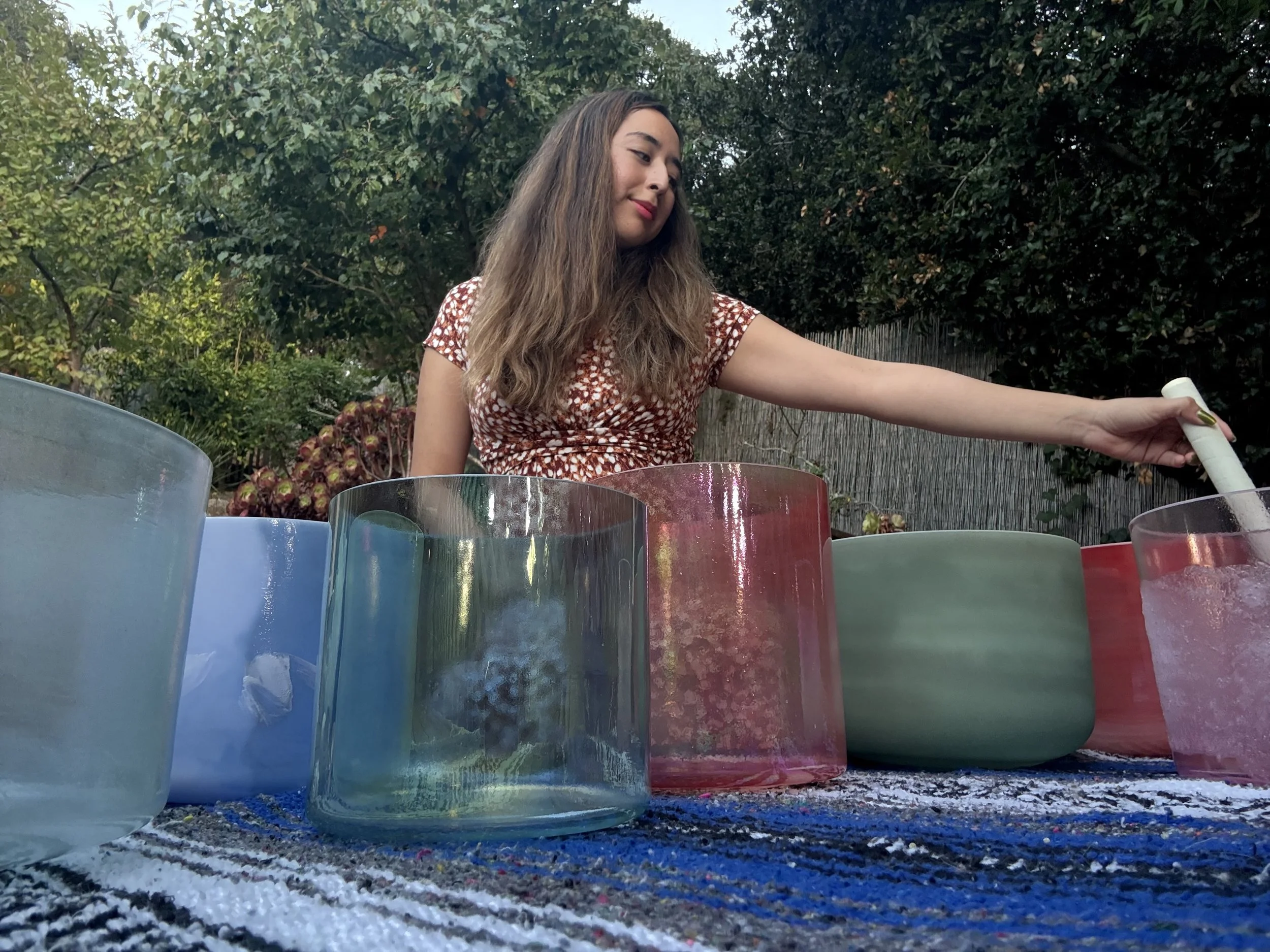 Young woman with long brown hair and red lipstick playing colorful glass or acrylic percussion instruments outdoors with trees in the background.