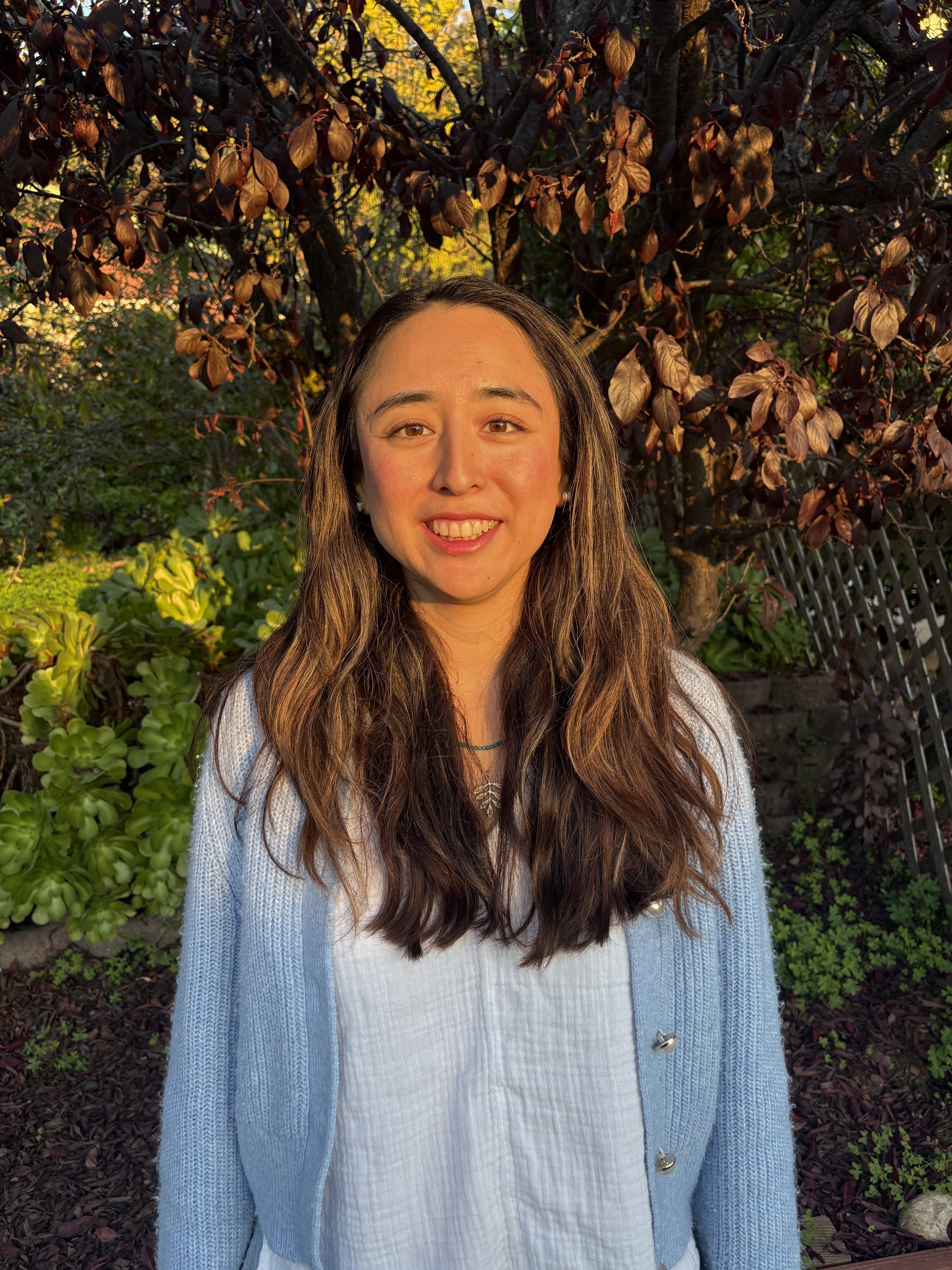 A young woman with long, wavy brown hair standing outdoors in front of a tree with dark purple leaves, smiling at the camera in warm, natural light.