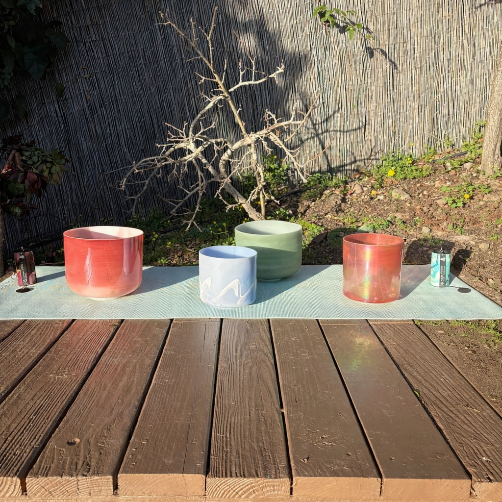 Five colorful singing bowls arranged on a light blue mat on a wooden deck outdoors, with a bamboo fence, plants, and a leafless tree in the background.