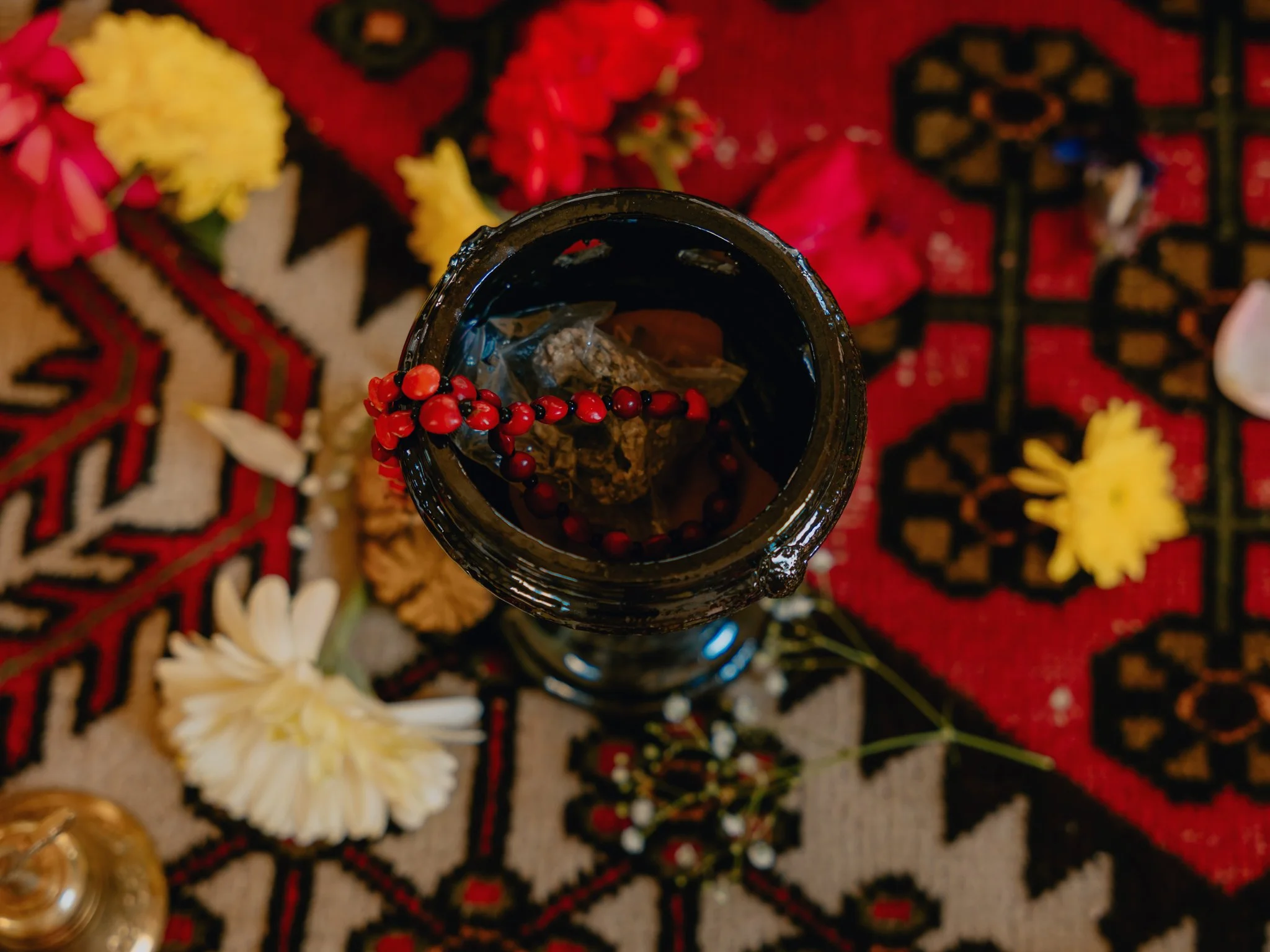 Top-down view of a dark glass jar on a patterned cloth, filled with water and botanical items, surrounded by flowers and decorative elements.
