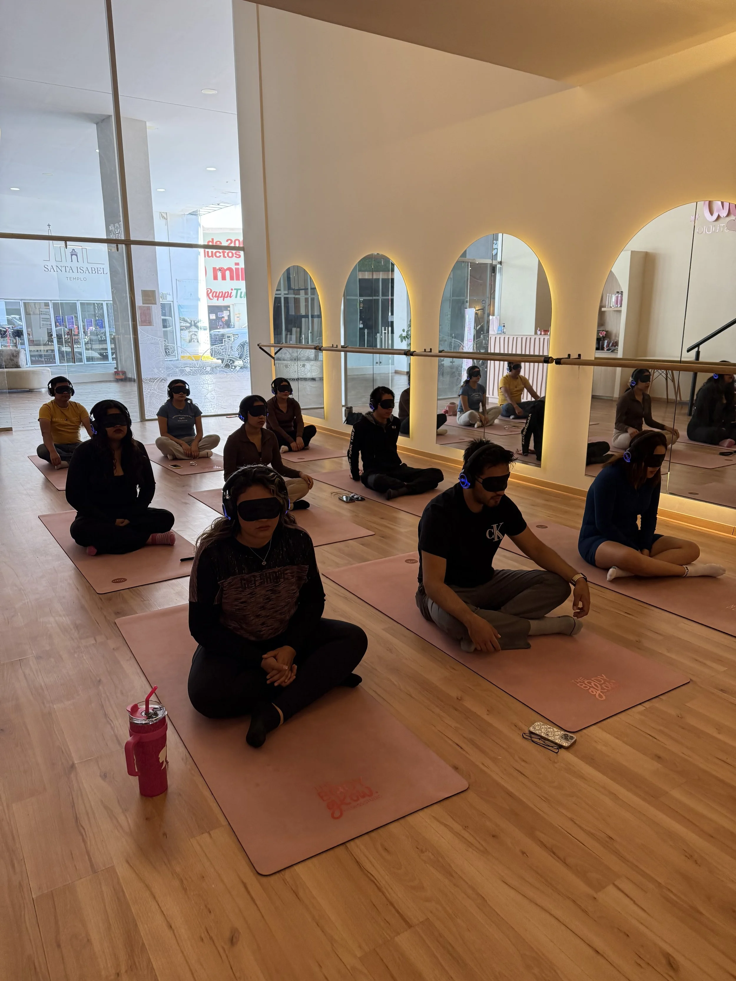 A group of people participating in a yoga or meditation class indoors, sitting cross-legged on pink mats, wearing eye masks, with a mirror along the wall reflecting their figures. Some have water bottles nearby, and there is a barre along the wall.