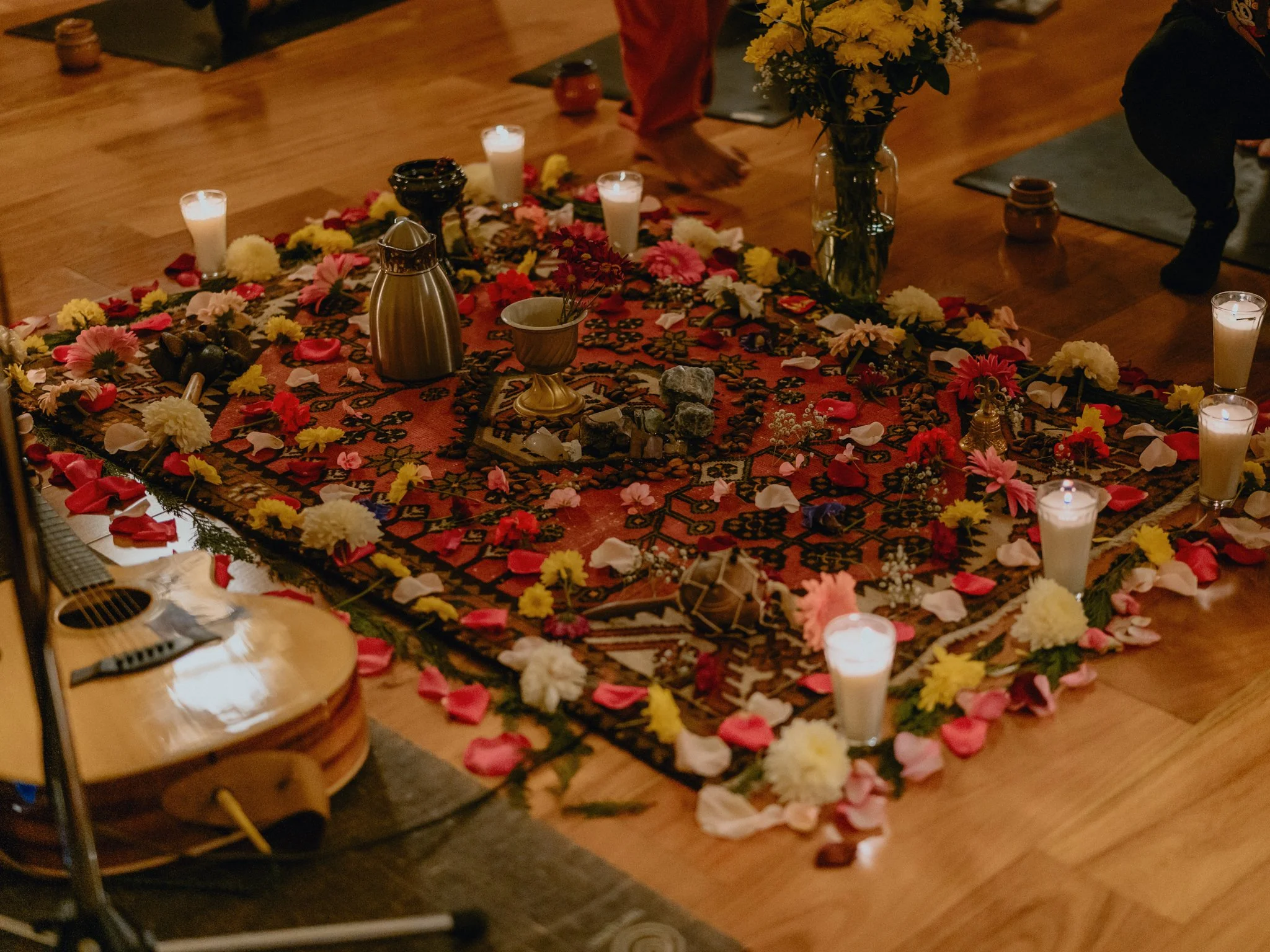 A decorated prayer or meditation space featuring a colorful patterned rug, surrounded by lit candles, flowers, and small ritual objects on a wooden floor, with people in the background.