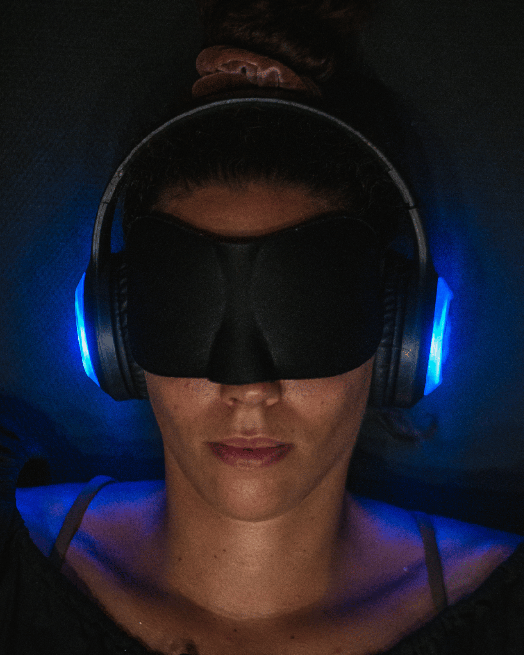 A woman lying down with a black sleep mask over her eyes, wearing illuminated blue headphones, and her hair tied back.