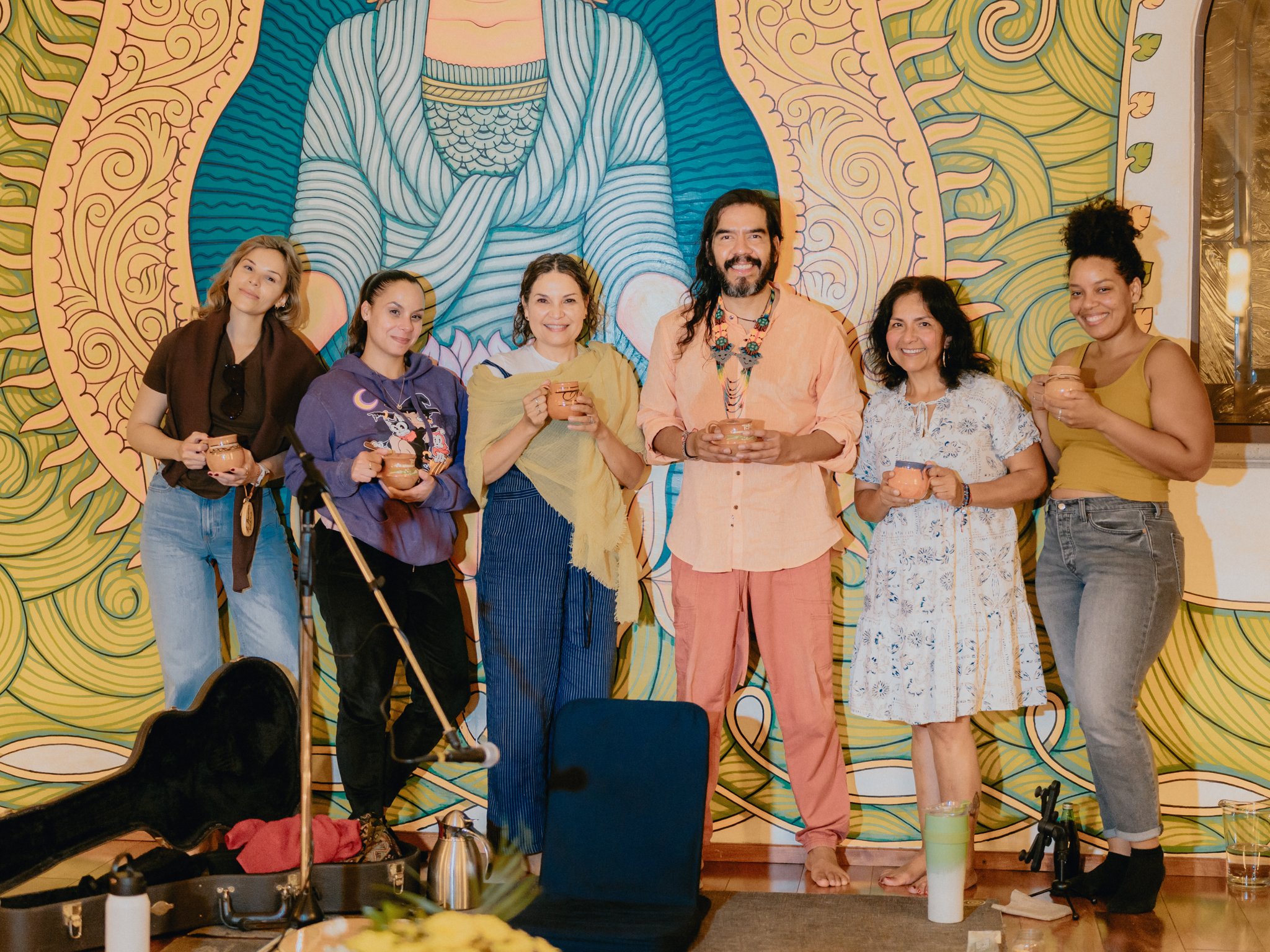 Group of seven people standing in front of a colorful mural, holding cups, and smiling at a gathering or celebration.