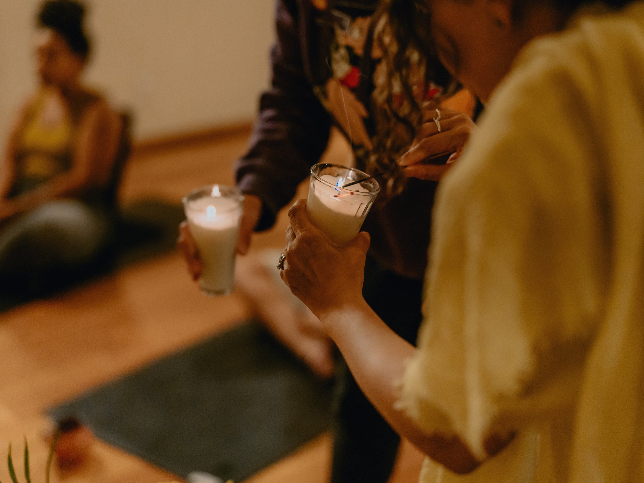 Two women holding lit candles in glasses during a gathering, with others seated in the background.