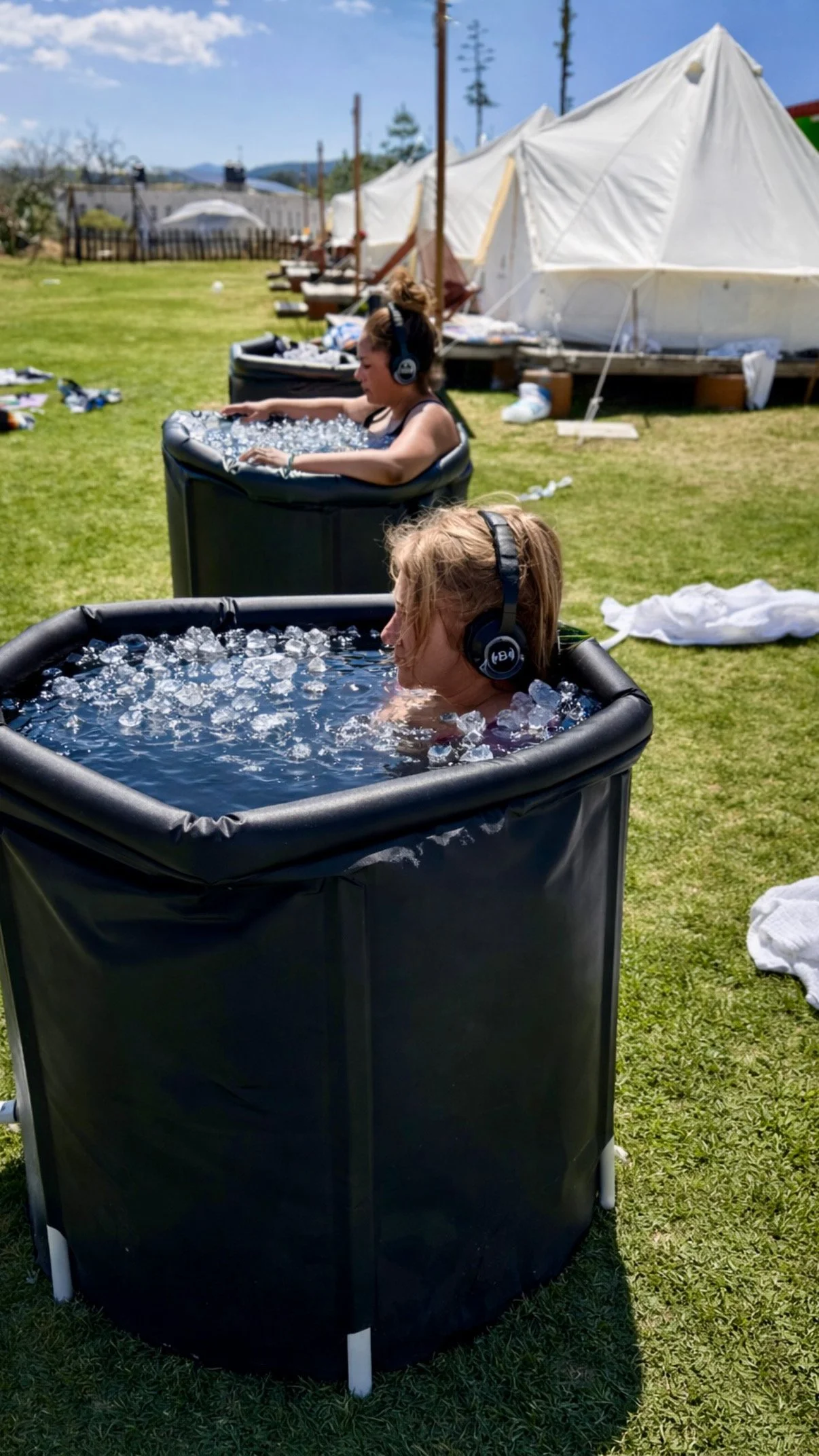 Two children wearing headphones sit in black inflatable tubs filled with water and ice outdoors on a sunny day, with white tents in the background.
