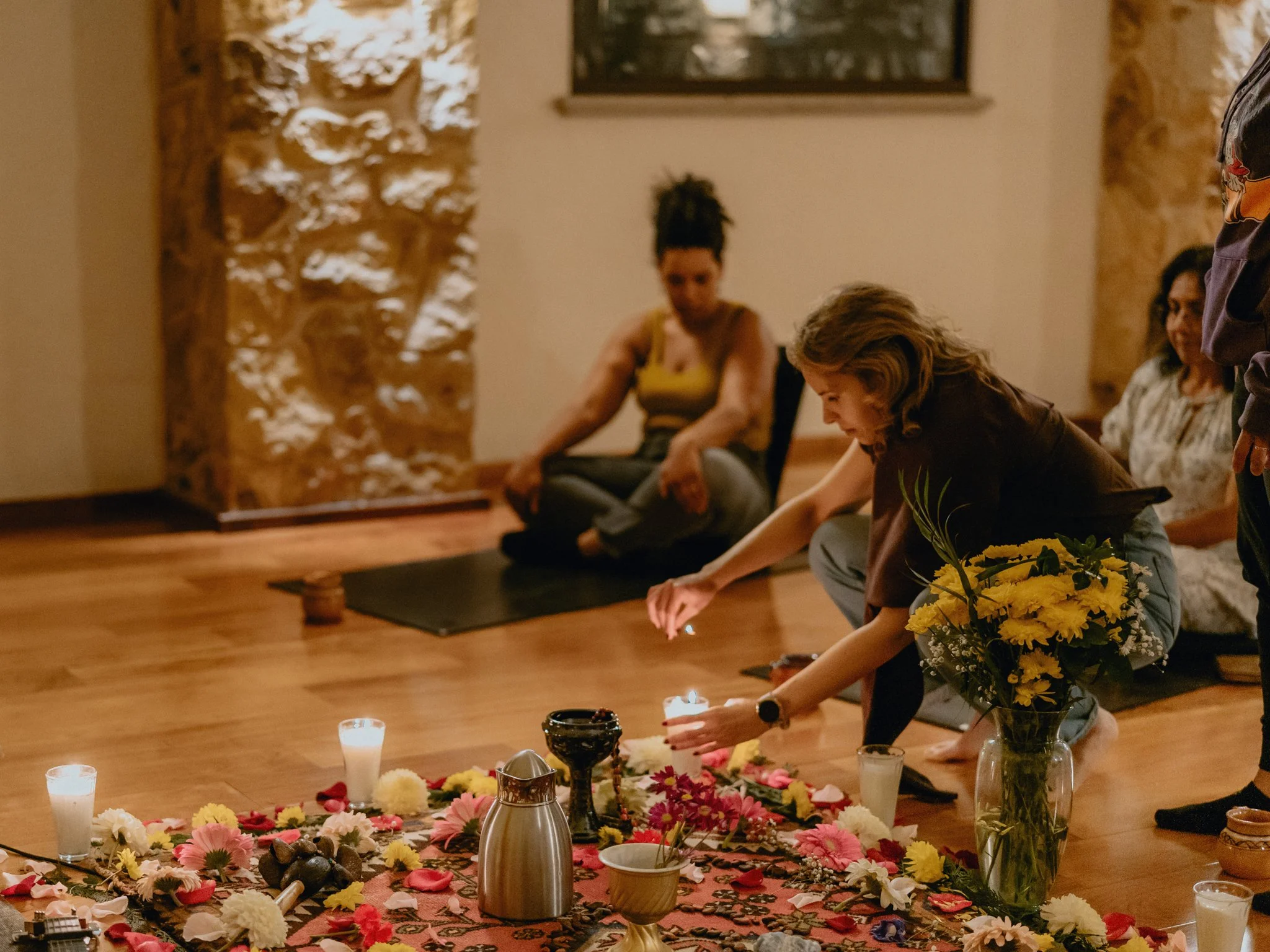 People participating in a meditation or prayer circle inside a room with wooden floors and stone walls. They are sitting on the floor, some with eyes closed, and a woman is lighting candles in front of a colorful floral arrangement on a decorative rug.