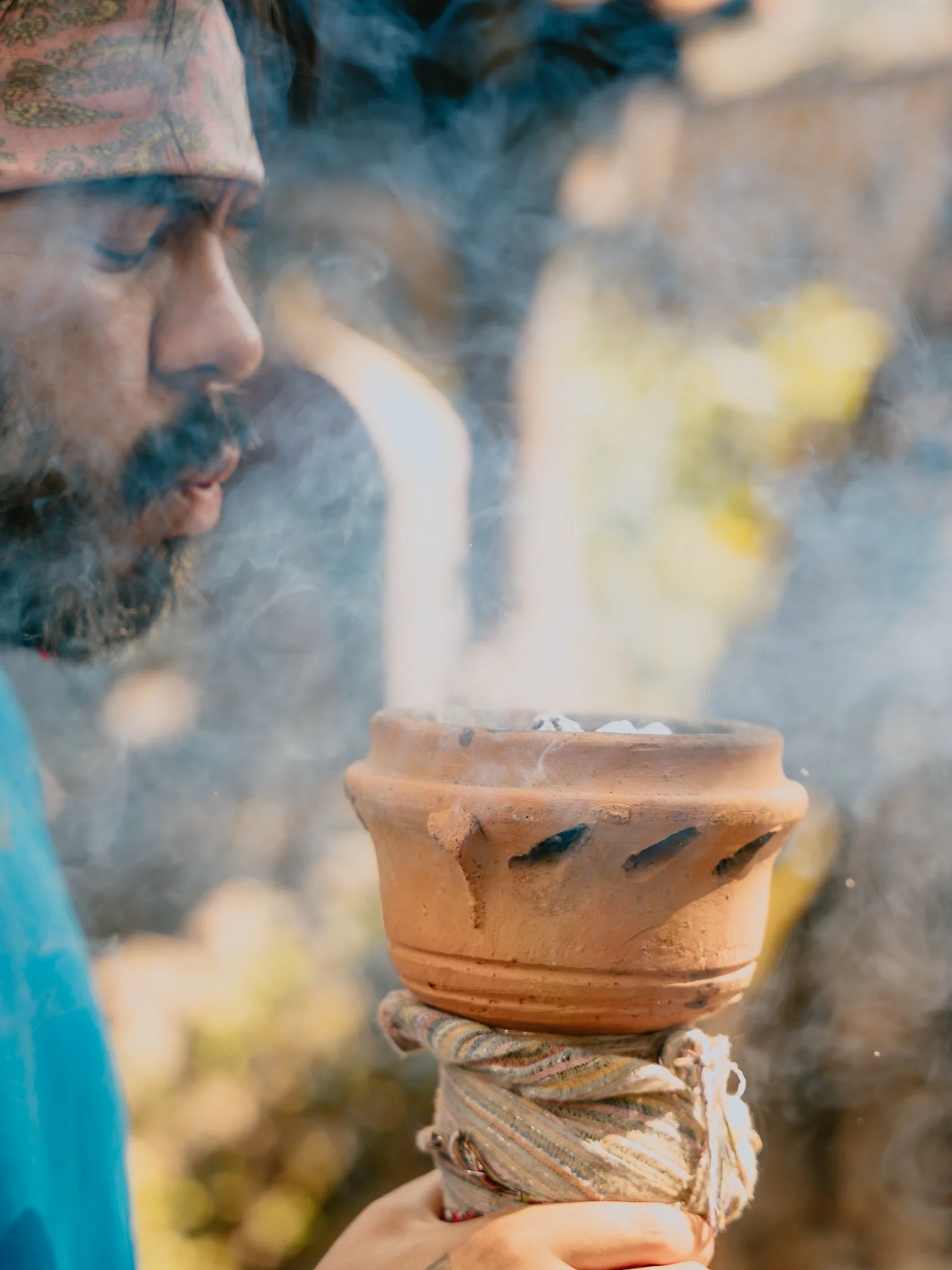 A man wearing a patterned headband blows smoke into the air while holding a traditional clay pipe with smoke rising from it.