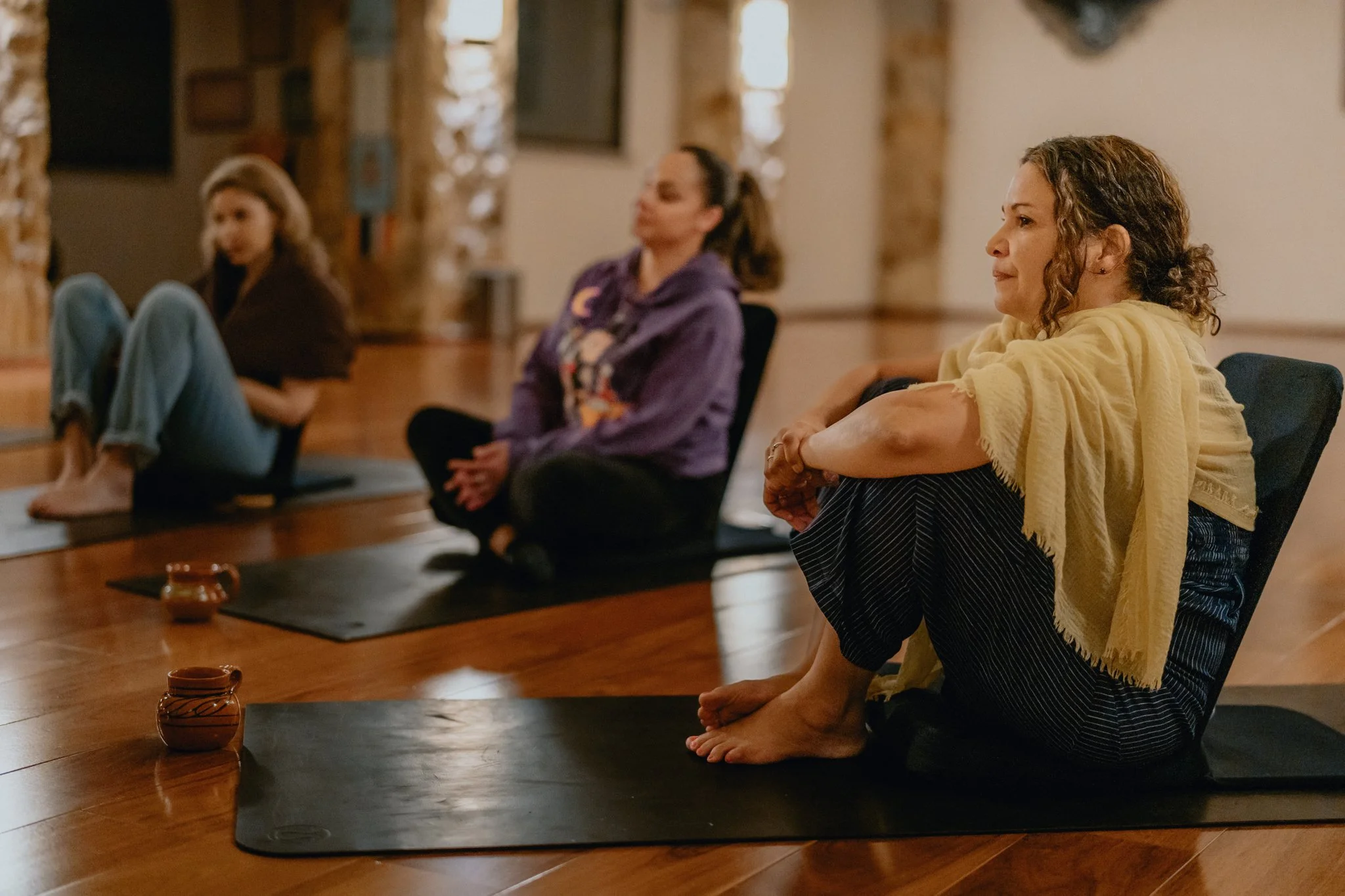 Three women participating in a yoga or meditation class sitting cross-legged on mats inside a room with wooden floors and brick walls.