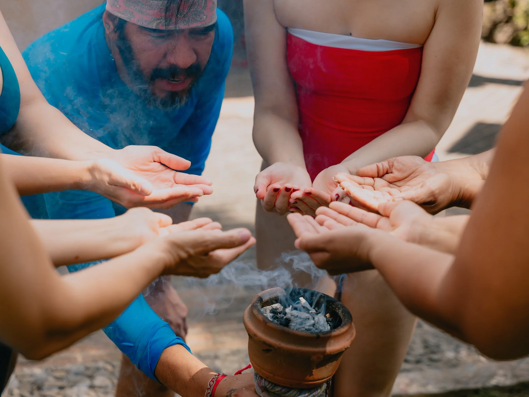 Group of people participating in a traditional outdoor ceremony, holding hands over a small fire in a clay pot.