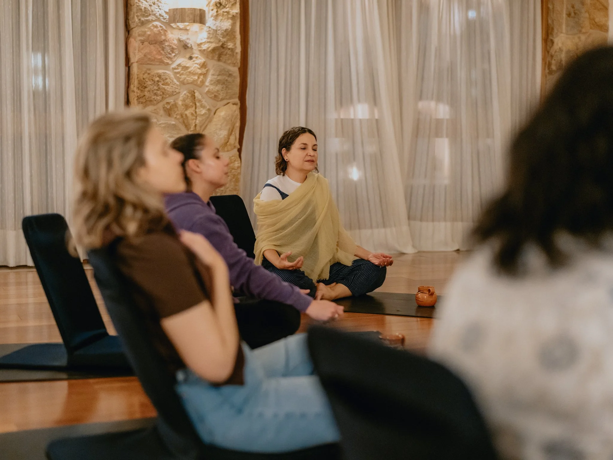 Women participating in a meditation session in a cozy room with stone walls and curtains, seated on mats and chairs.