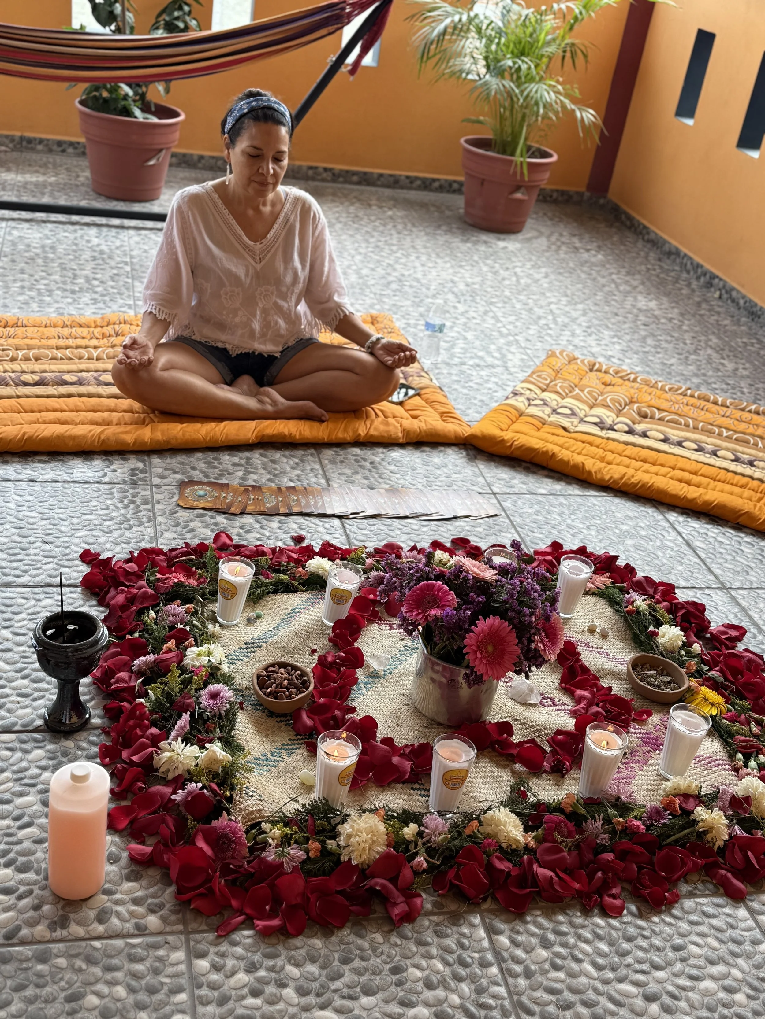 A woman sitting cross-legged on a blanket, meditating in front of a floral altar with candles, petals, and a vase of flowers.