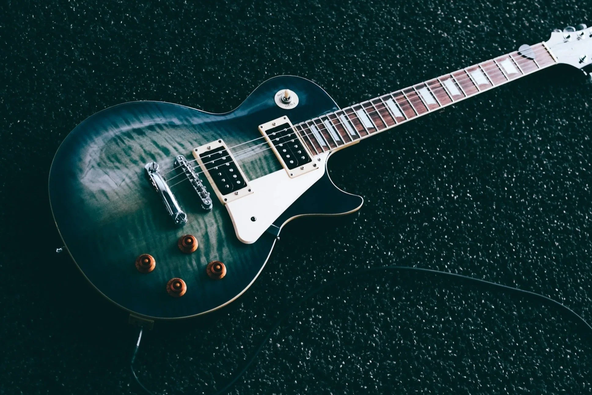 A blue electric guitar with a flamed maple top, white pickguard, two humbucker pickups, four orange control knobs, and a rosewood fretboard on a black textured surface.