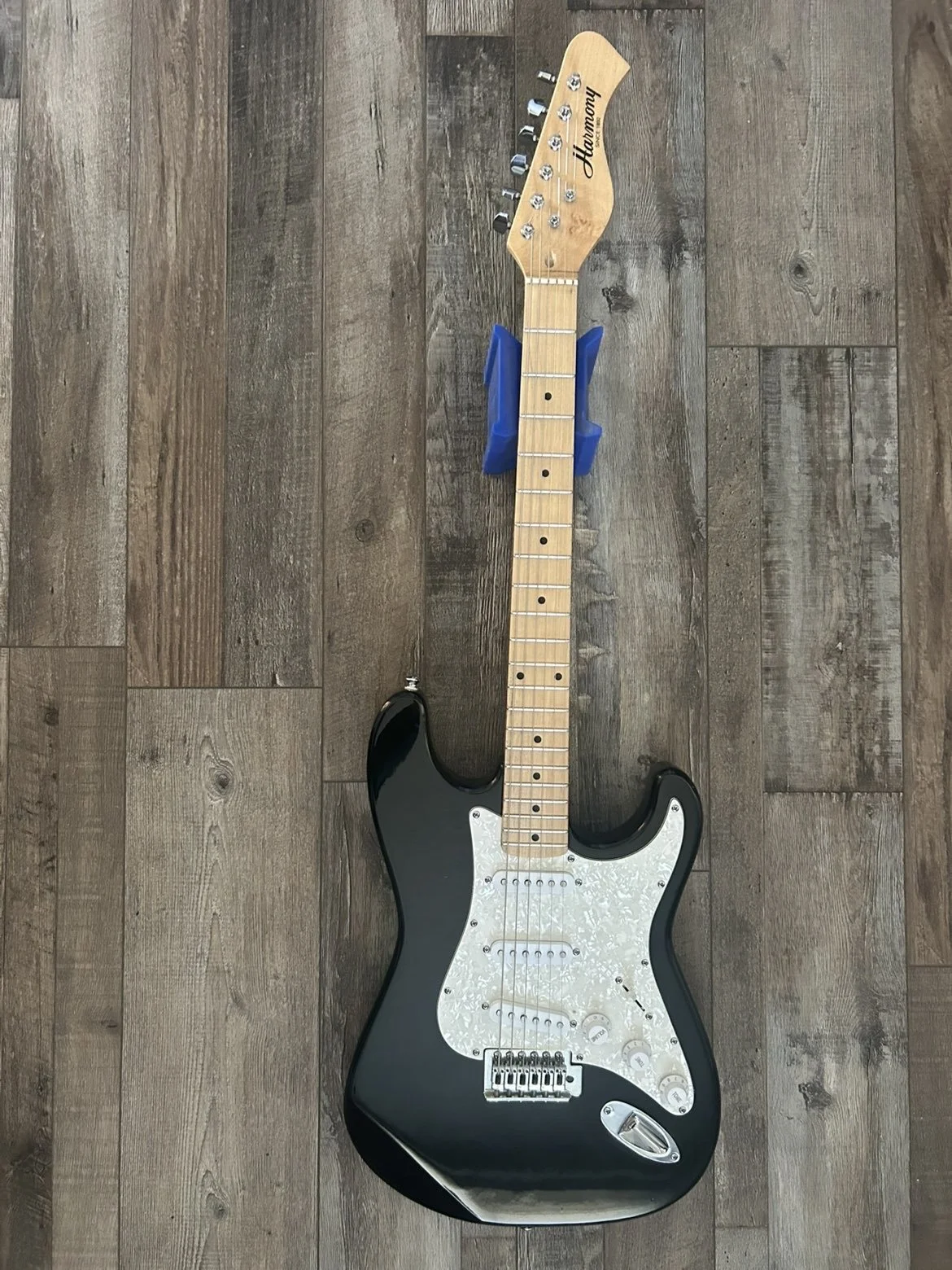 Black electric guitar with a white pickguard, mounted on a wooden floor with a blue guitar stand behind the neck.