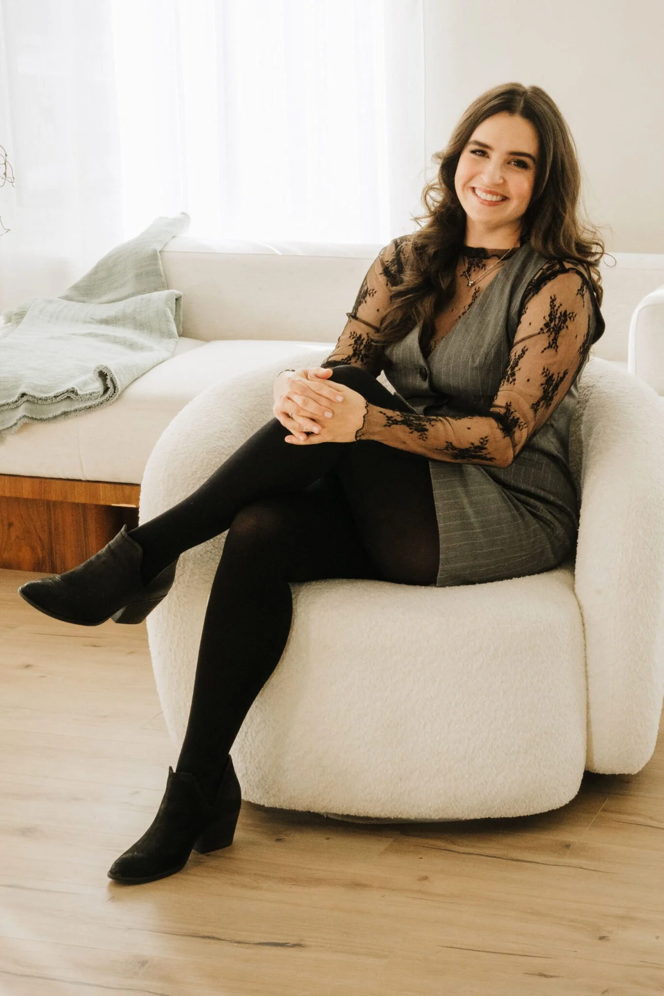 Samantha Heuwagen, Licensed Marriage and Family Therapist, sitting in a white armchair smiling in a grey pinstriped dress with black lace sleeves and black tights