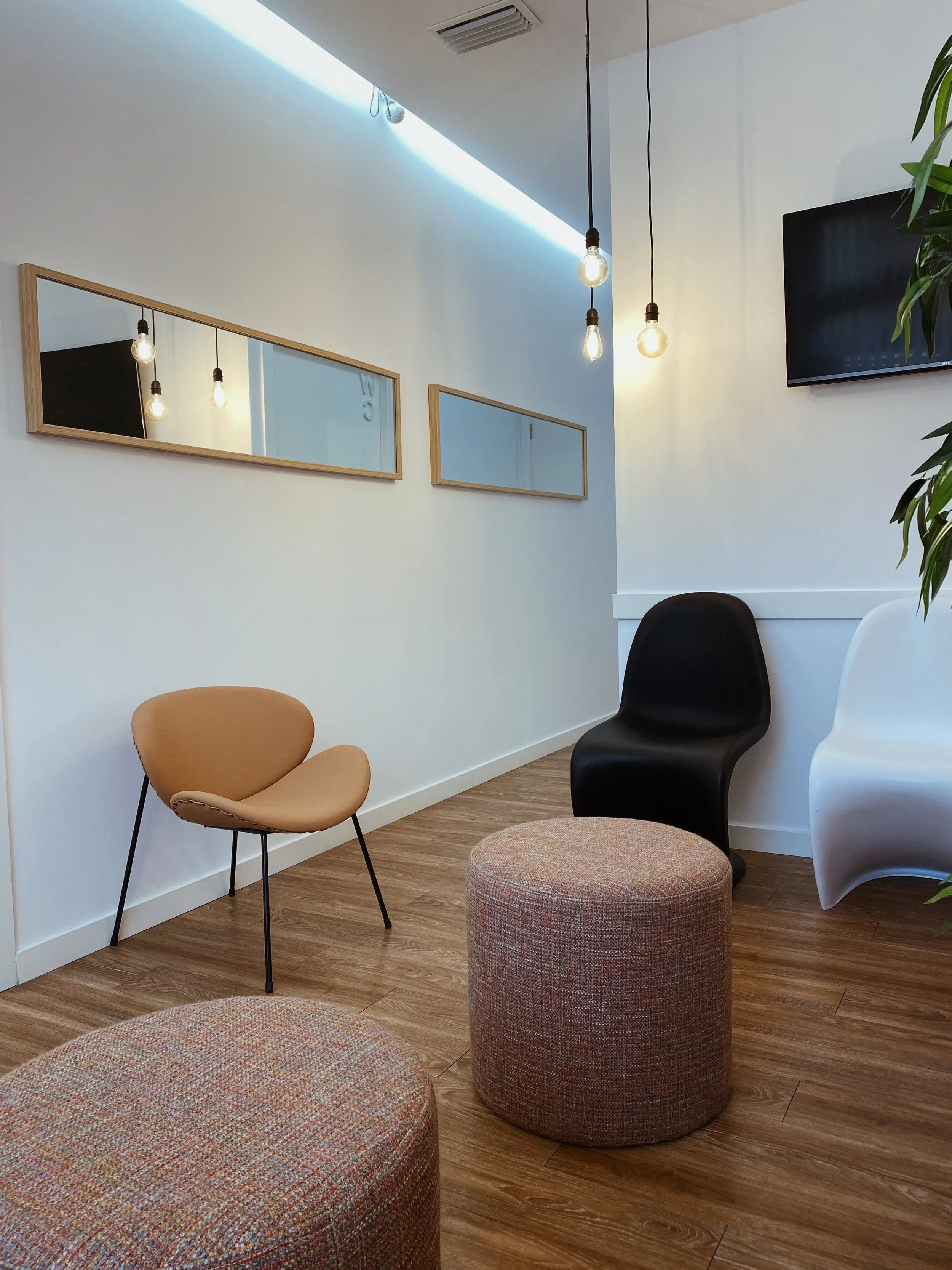 A modern interior with white walls, wooden flooring, and hanging light bulbs. There are two oval mirrors on the wall, a black TV mounted on the wall, and various chairs including a tan Chair, a black chair, and a white chair. A green plant is partially visible on the right.