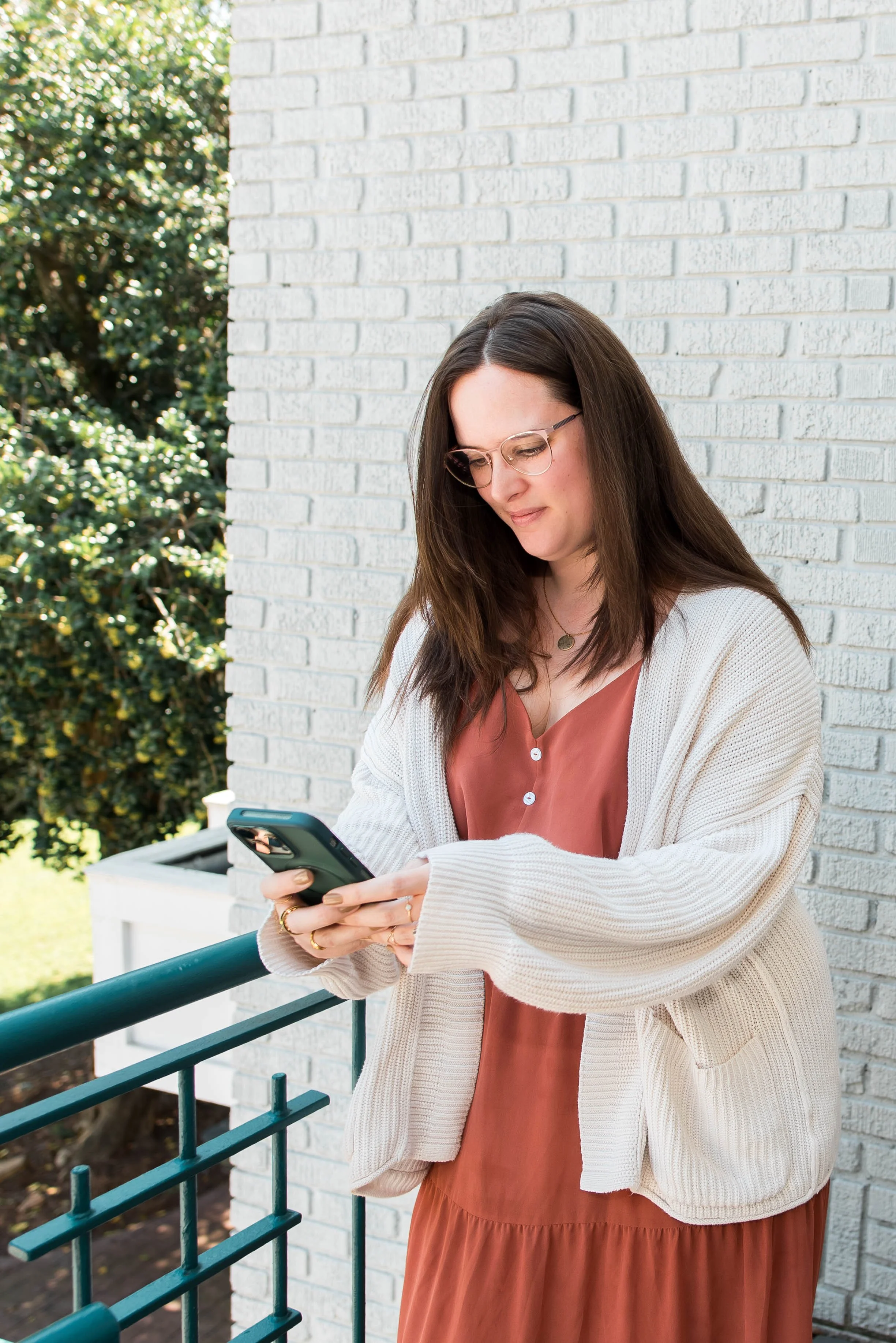 A woman with long brown hair and glasses is standing outdoors on a porch, looking at her smartphone, with a white brick wall and green bushes in the background.