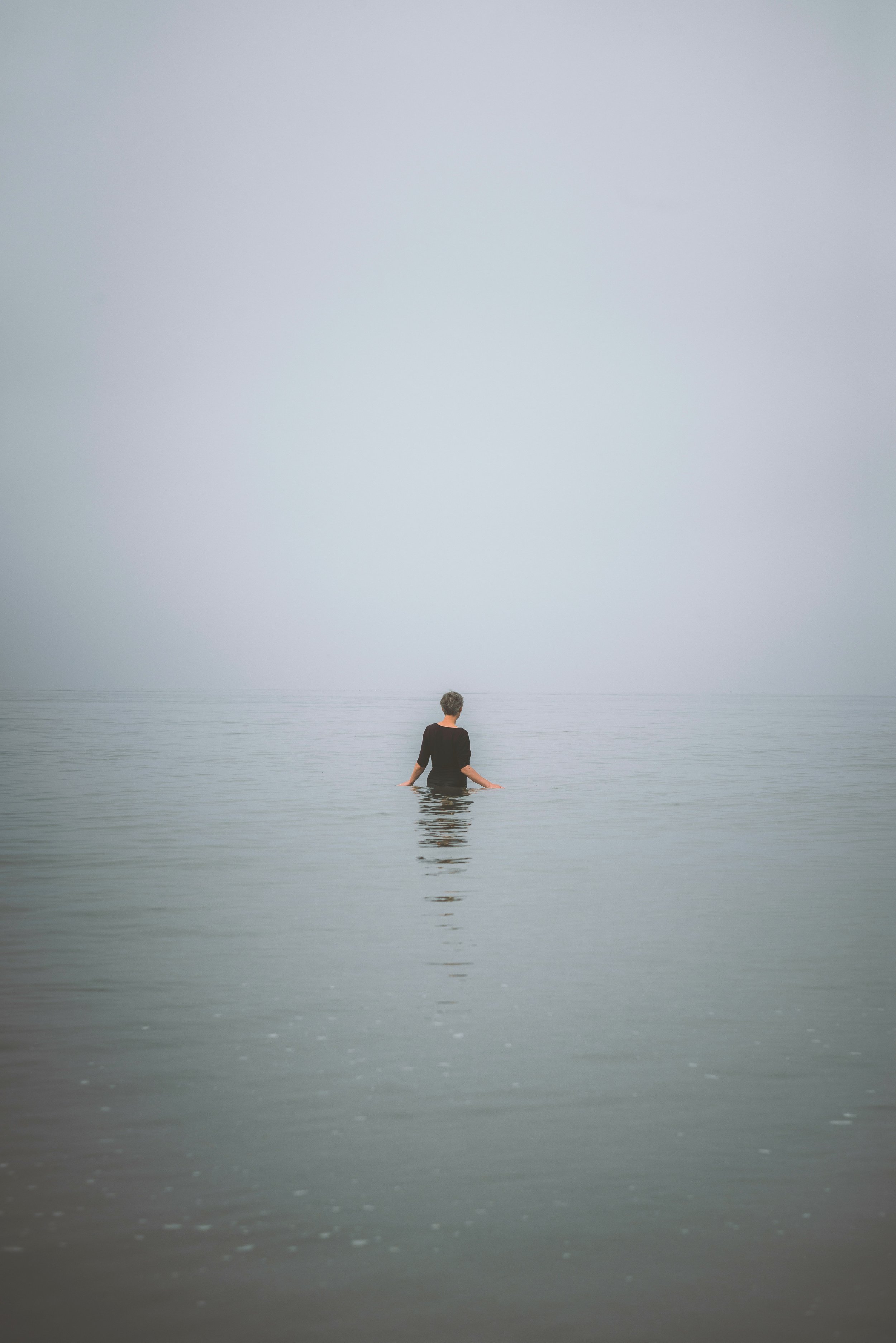 A person with short hair, dressed in black, standing in calm water up to their waist, looking out into a foggy, grey horizon.