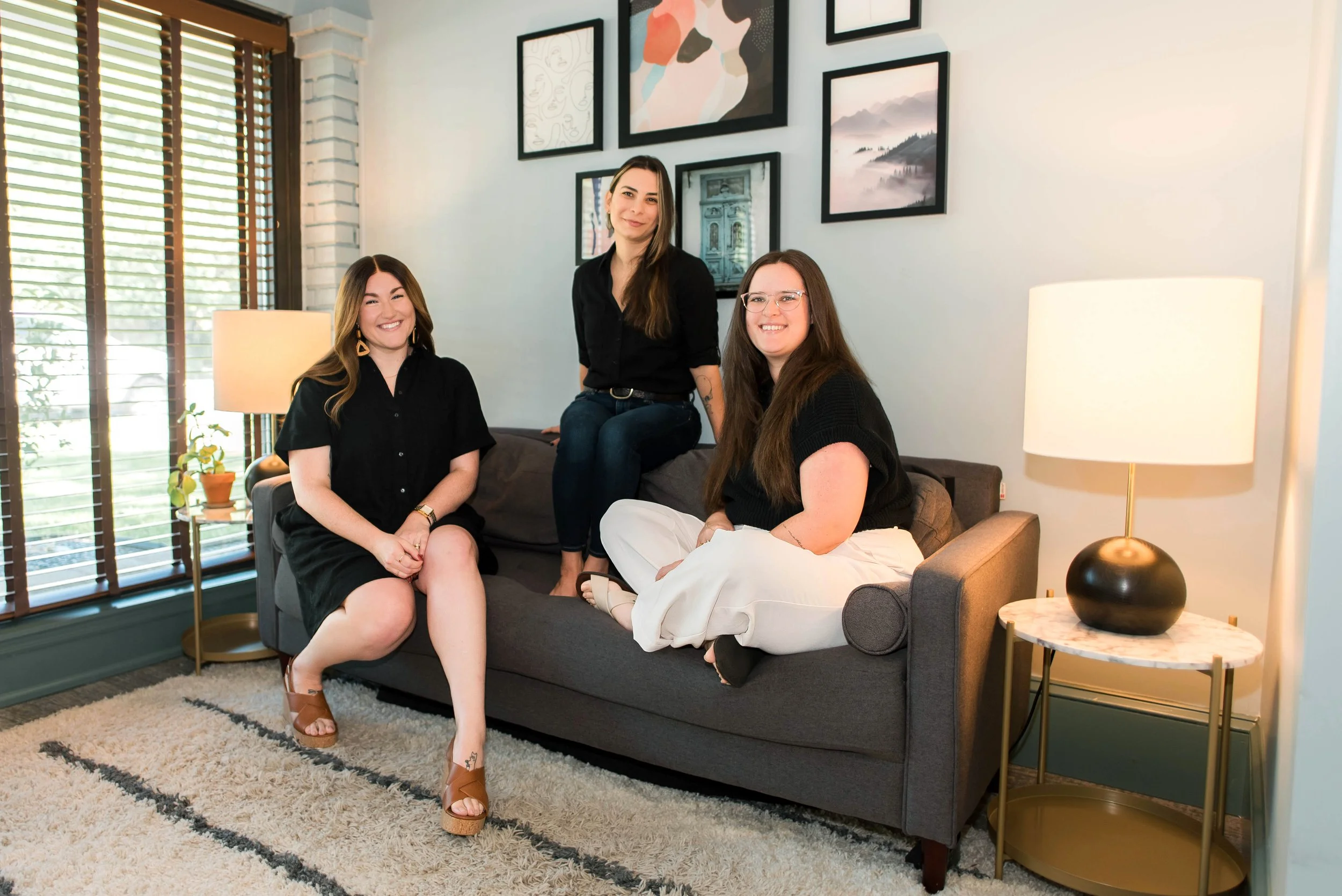 Four women sitting and standing on a gray sofa in a modern living room with framed wall art, a window with blinds, and a side table with a lamp.