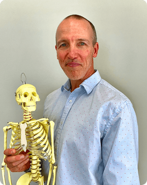 Rick Olderman, physical therapist, smiling wearing a light blue button-down shirt while holding a small anatomical model of a human skeleton