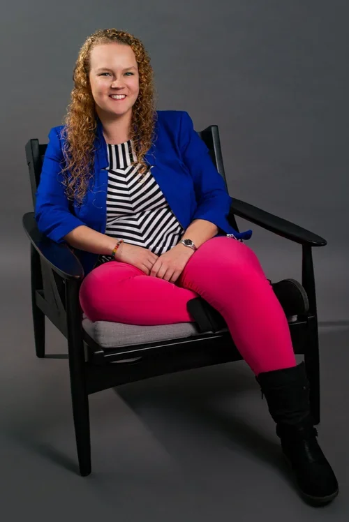 Karen Lill, Licensed Professional Counselor, smiling in a bright blue blazer and hot pink pants while sitting in an armchair against a gray backdrop