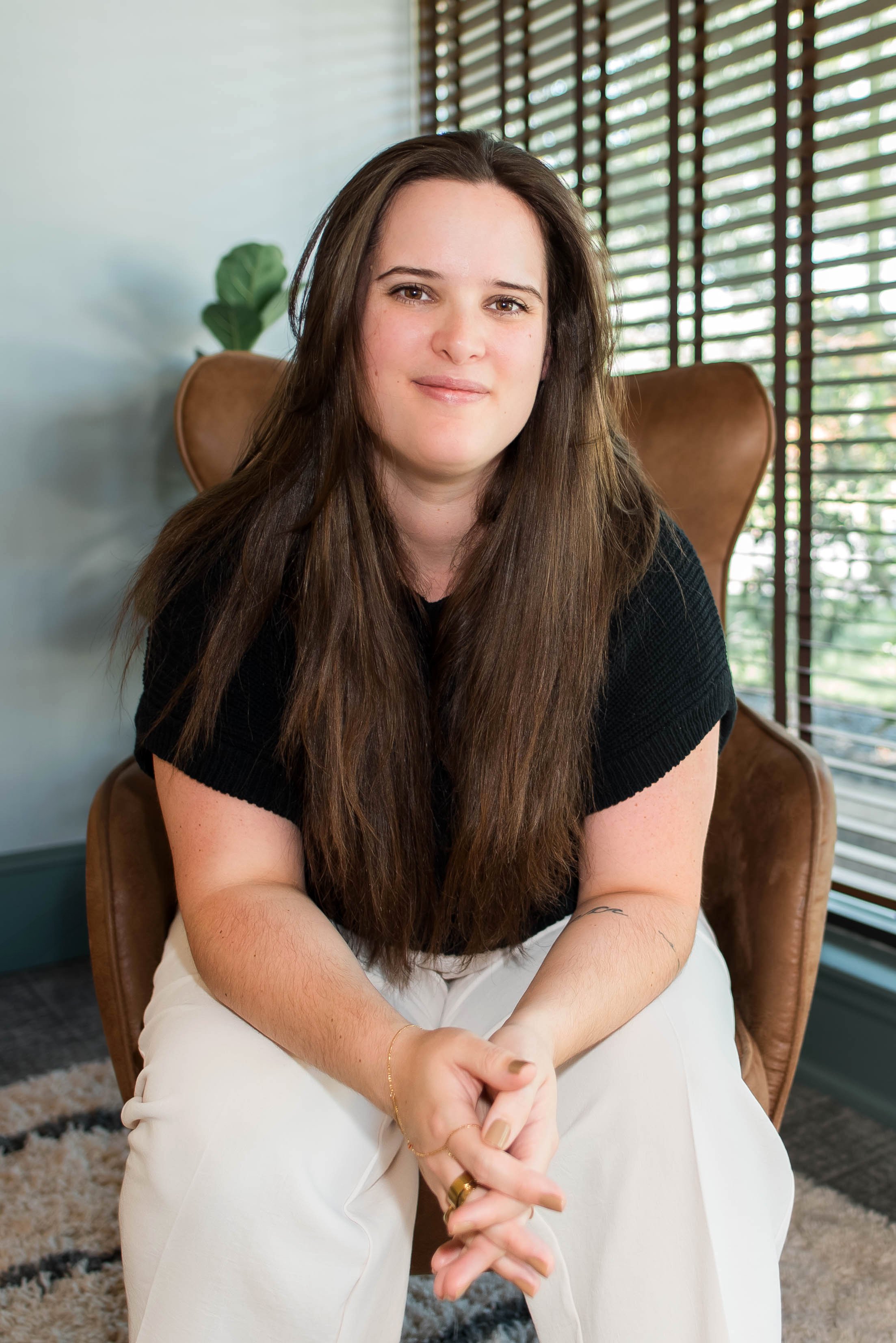 A woman with long brown hair sitting in a leather chair in front of a window with wooden blinds, wearing a black top and light-colored pants.