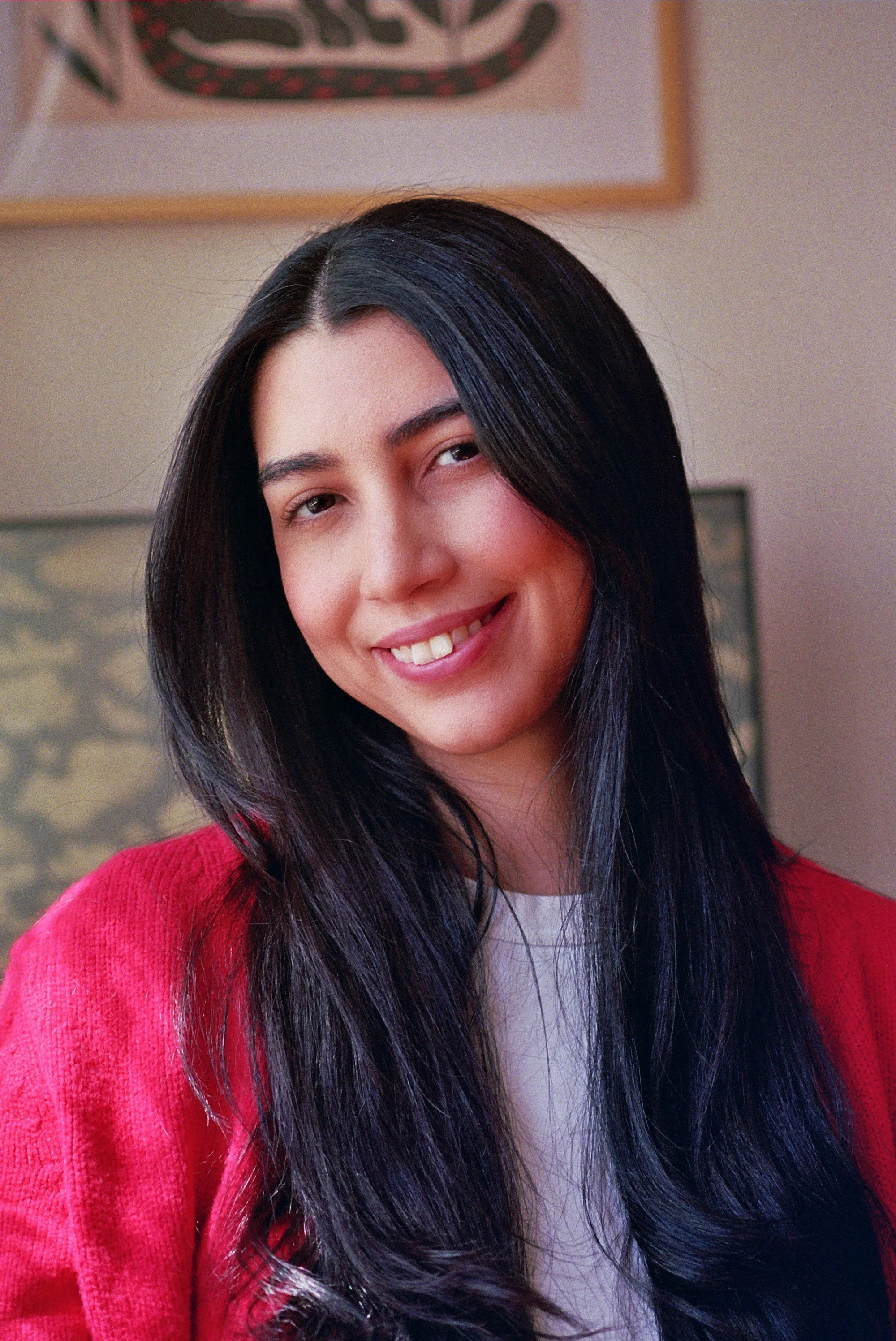 A woman with long black hair, smiling, wearing a red sweater over a white top, standing indoors with framed art on the wall behind her.