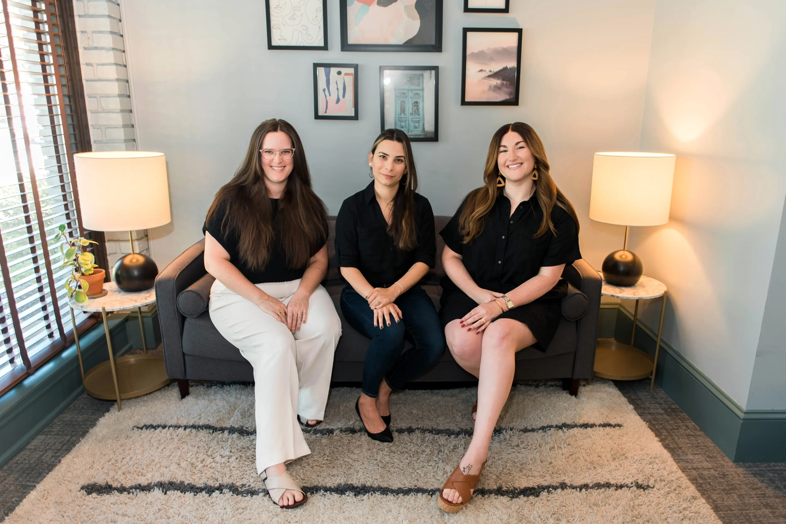 Three women sitting on a gray sofa in a living room, smiling at the camera. The woman on the left has long brown hair, glasses, a black top, and white pants. The woman in the middle has shoulder-length dark hair, earrings, a black shirt, and dark jeans. The woman on the right has long wavy brown hair, earrings, a black dress, and brown sandals. Behind them, framed artwork and photos are displayed on a white wall. Two lamps with beige shades illuminate the room, which features a window with wooden blinds, a potted plant on a side table, a cream-colored rug, and blue-green walls.