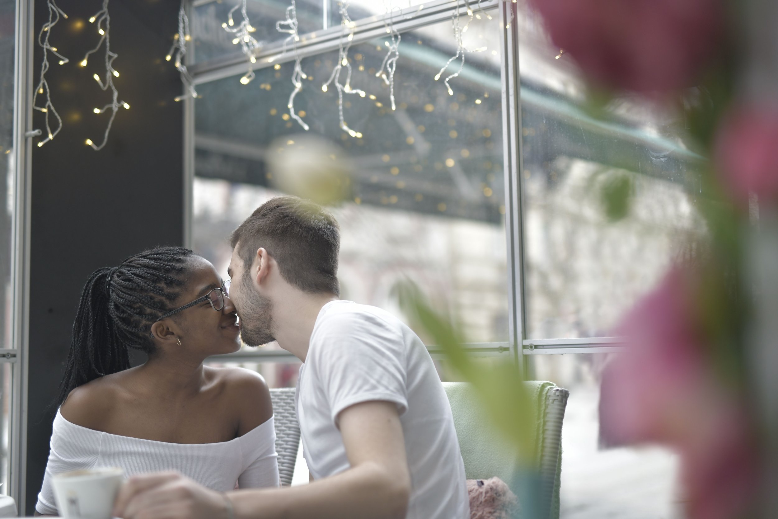 A couple leaning in for a kiss while sitting close together, with string lights hanging overhead and blurred plants surrounding them.