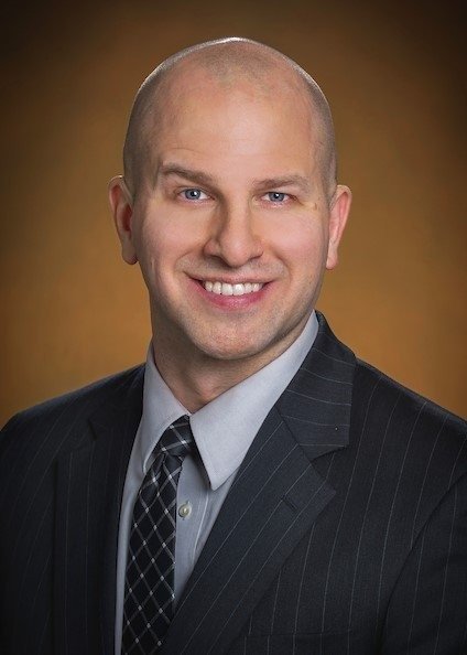 Mark Kargela, physical therapist and founder, smiling in a dark pinstripe suit jacket, light gray dress shirt, and patterned tie against a warm gold studio background
