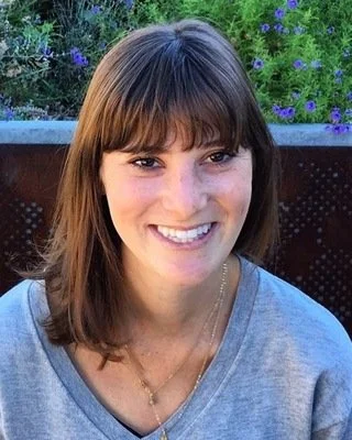 Zara Drapkin, Licensed Clinical Social Worker, smiling in a grey v-neck top and delicate layered gold necklaces outdoors