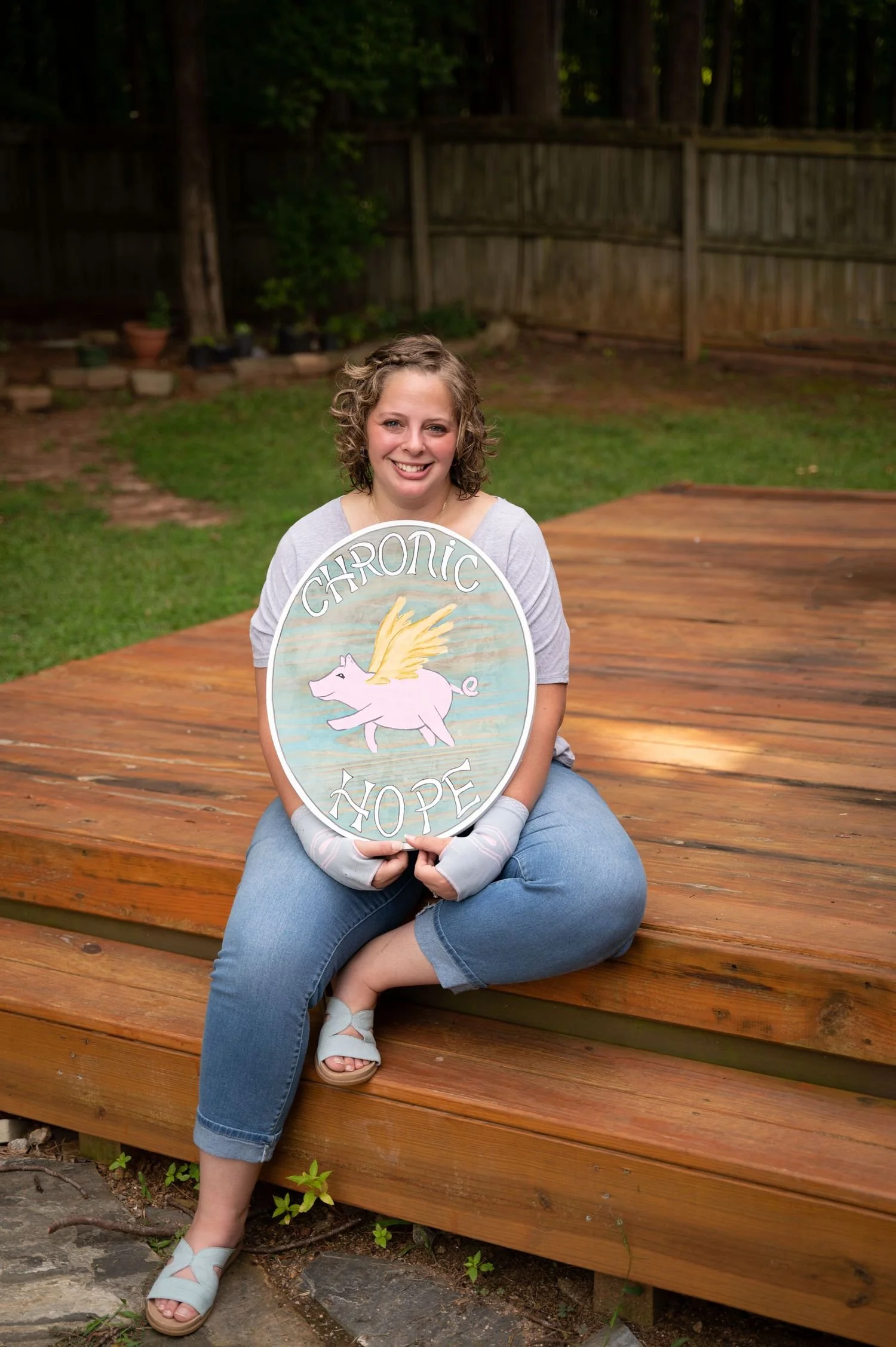 Jennifer Sutton, Licensed Clinical Mental Health Counselor Associate and founder, smiling in a grey top, cuffed jeans, and wrist braces while holding an oval sign reading "CHRONIC HOPE"