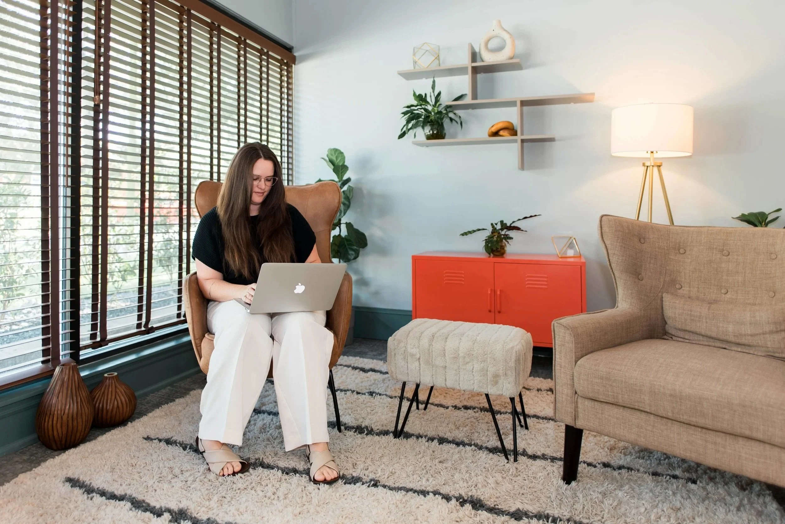 A woman with long brown hair, glasses, black top, and white pants sitting in a beige armchair in a living room, working on a silver laptop. The room has large windows with wooden blinds, a beige couch, a white lampshade on a gold tripod lamp, green plants, and decorative shelves with art objects and a banana. The floor is covered with a patterned rug.