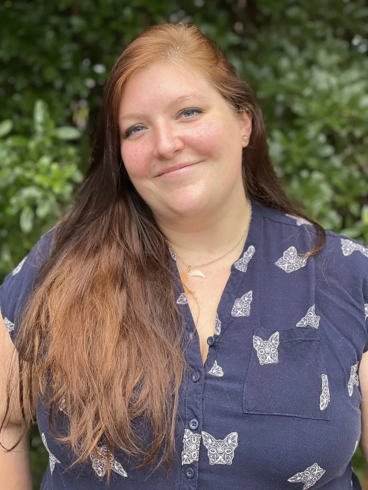Rowan Crawfish, counselor and art therapist, smiling wearing a navy printed shirt in front of green foliage