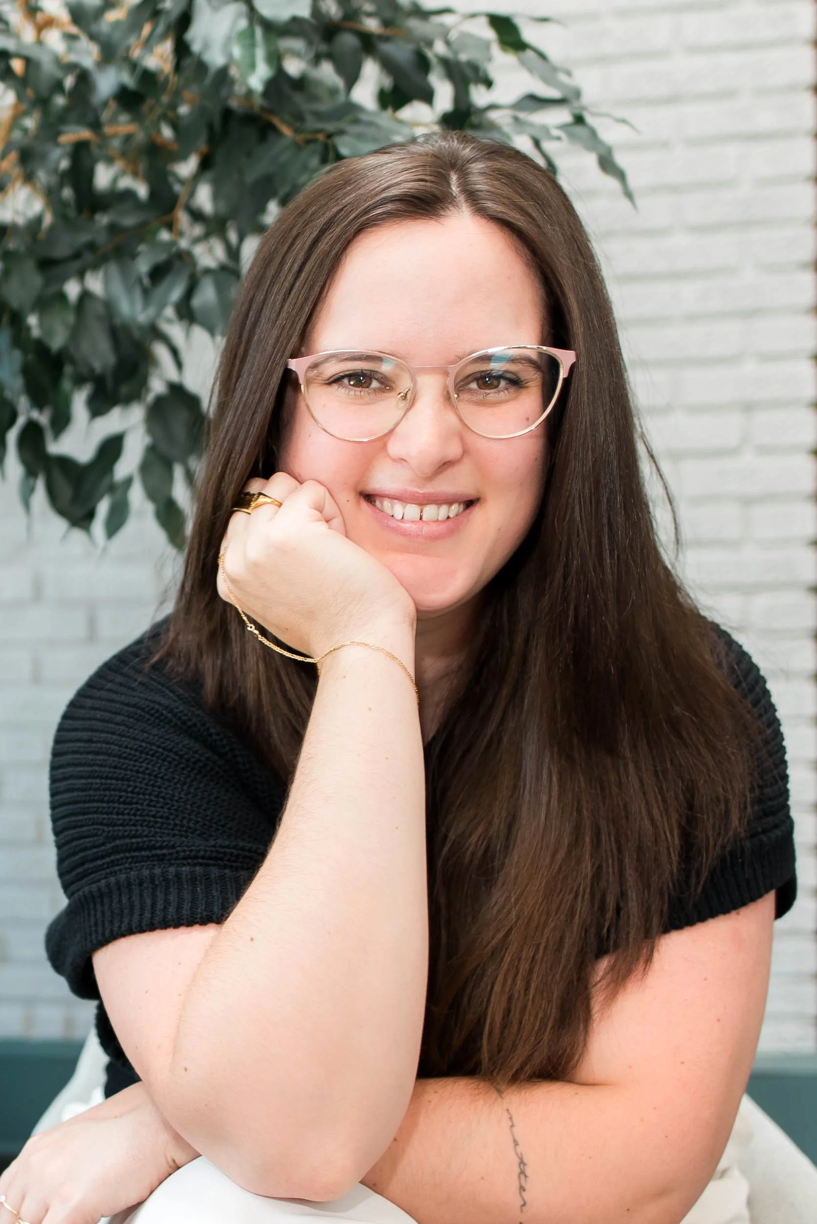 Close-up of a smiling woman with long brown hair, wearing glasses and a black top, sitting with her left arm resting on her knee, in front of a leafy plant and a white brick wall.