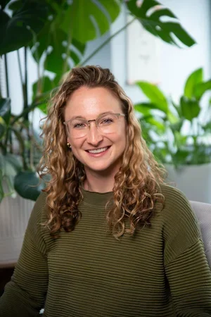 Samantha Allweiss, Licensed Clinical Social Worker,  smiling wearing an olive green ribbed sweater, sitting in front of houseplants.