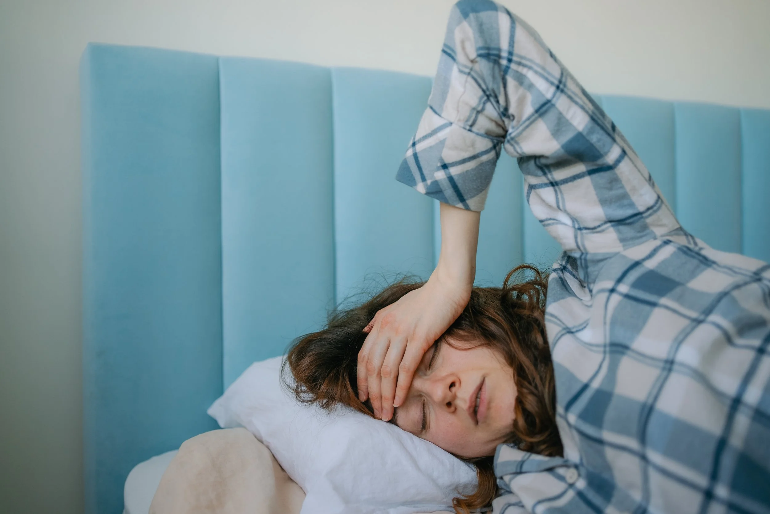 A woman with brown hair lying in bed with her eyes closed, holding her forehead with her left hand, appearing distressed. A child with curly hair, dressed in a plaid shirt, is leaning over her, touching her forehead with their right hand.