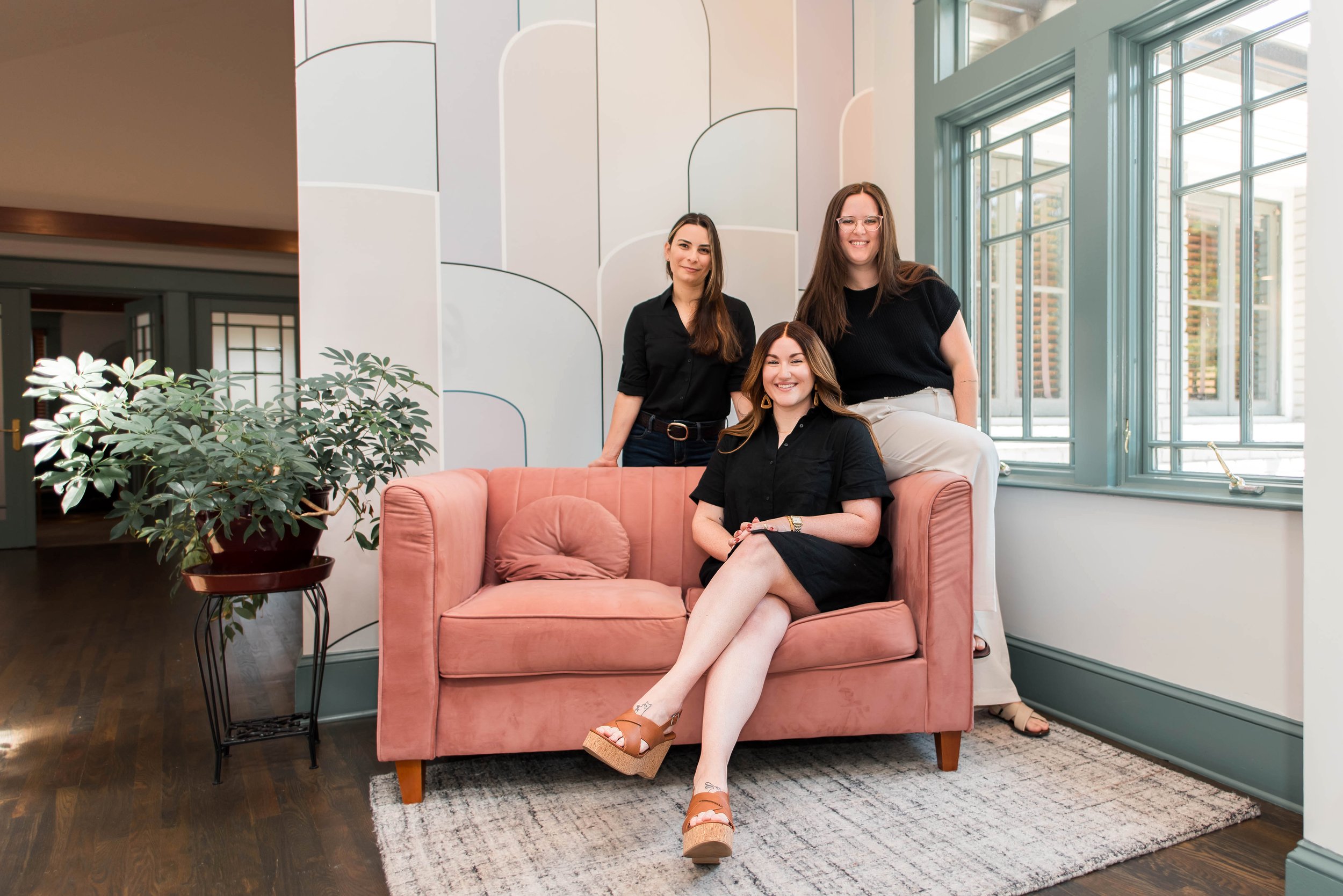 Three women posing inside a bright, modern room with large windows and colorful geometric wall art. One woman is sitting on a pink sofa, smiling, with crossed legs and wearing a black dress and tan wedge sandals. Two women are standing behind her, smiling, one with long brown hair in a black shirt and the other with long brown hair, glasses, and a black top with light-colored pants.