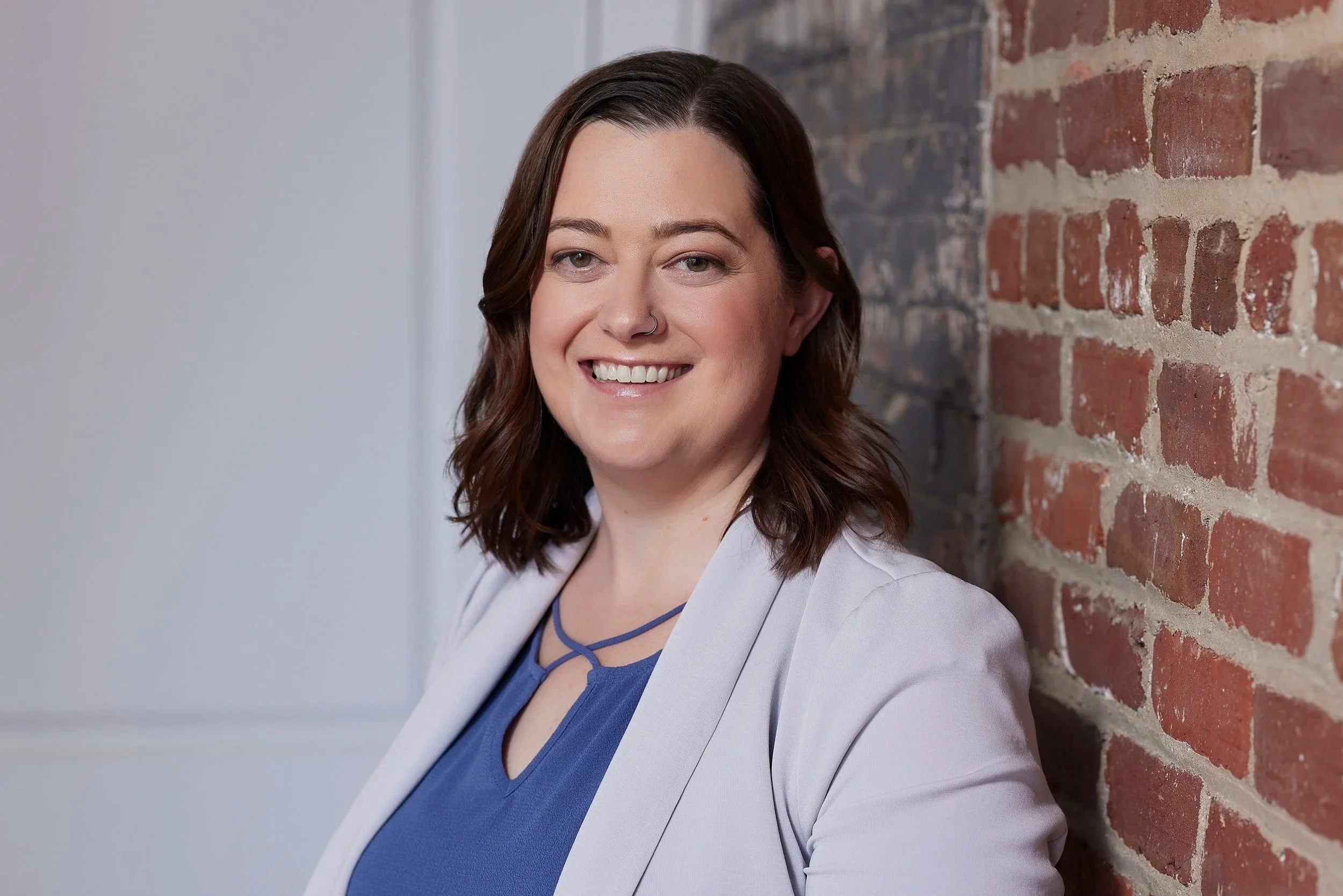 Dr. Ginny Kington, licensed psychologist, smiling in a light blazer and blue top leaning against an exposed red brick wall indoors