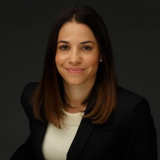 Dr. Ana Arteaga-Biggs, Licensed Clinical Psychologist, smiling in a black suit jacket and a white crewneck top against a dark backdrop