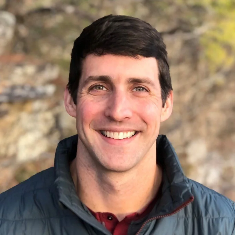 Jason Therrien, holistic physical therapist and health coach, smiling in a dark blue zip-up jacket over a maroon shirt against a soft-focus outdoor background