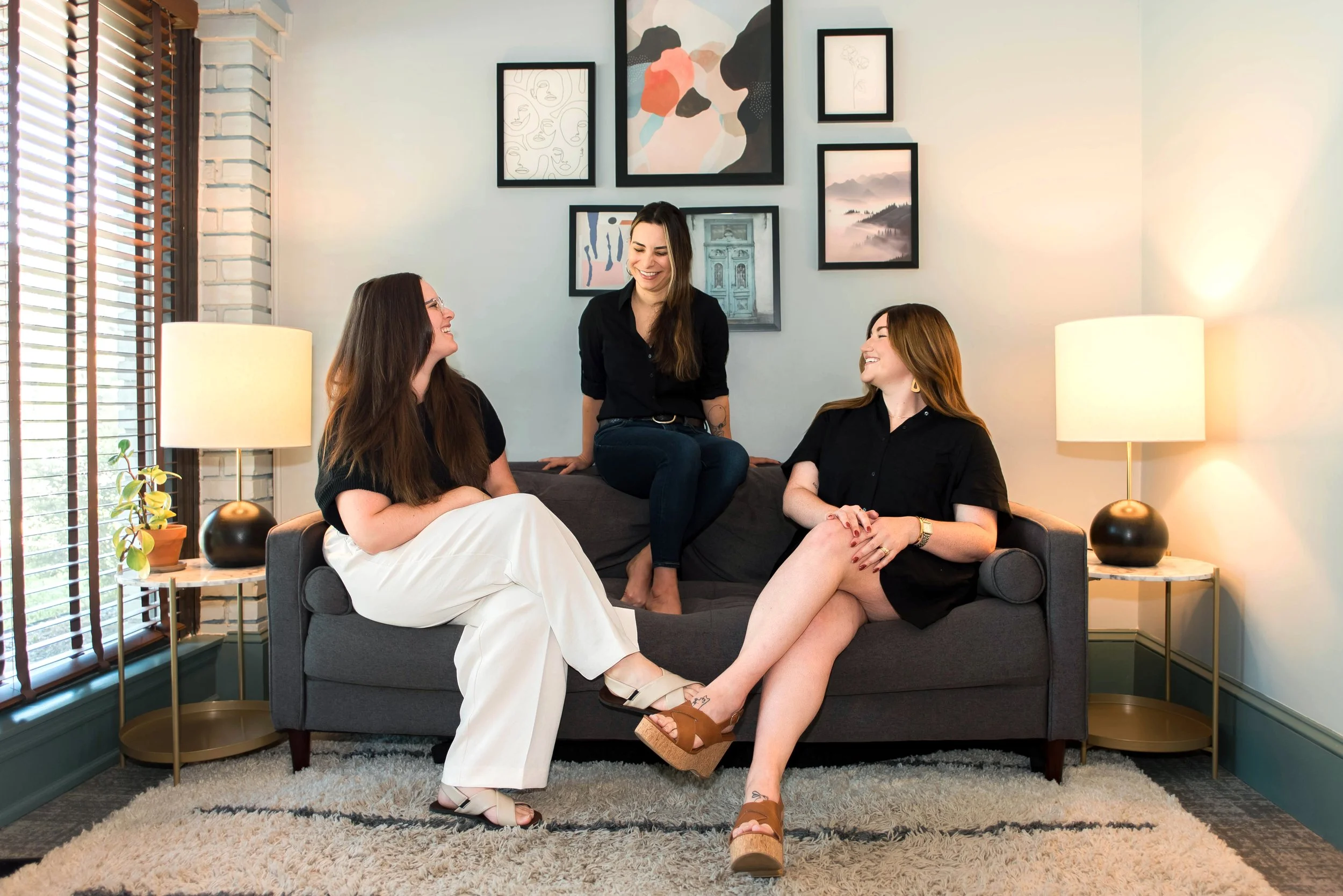 Three women having a friendly conversation in a cozy living room with artwork on the wall and warm lighting.