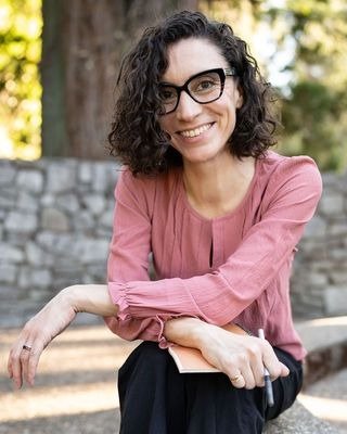 Candice Craft, Licensed Marriage and Family Therapist, smiling in a dusty pink long-sleeve blouse, resting her arms on her lap while holding a small notebook and pen.
